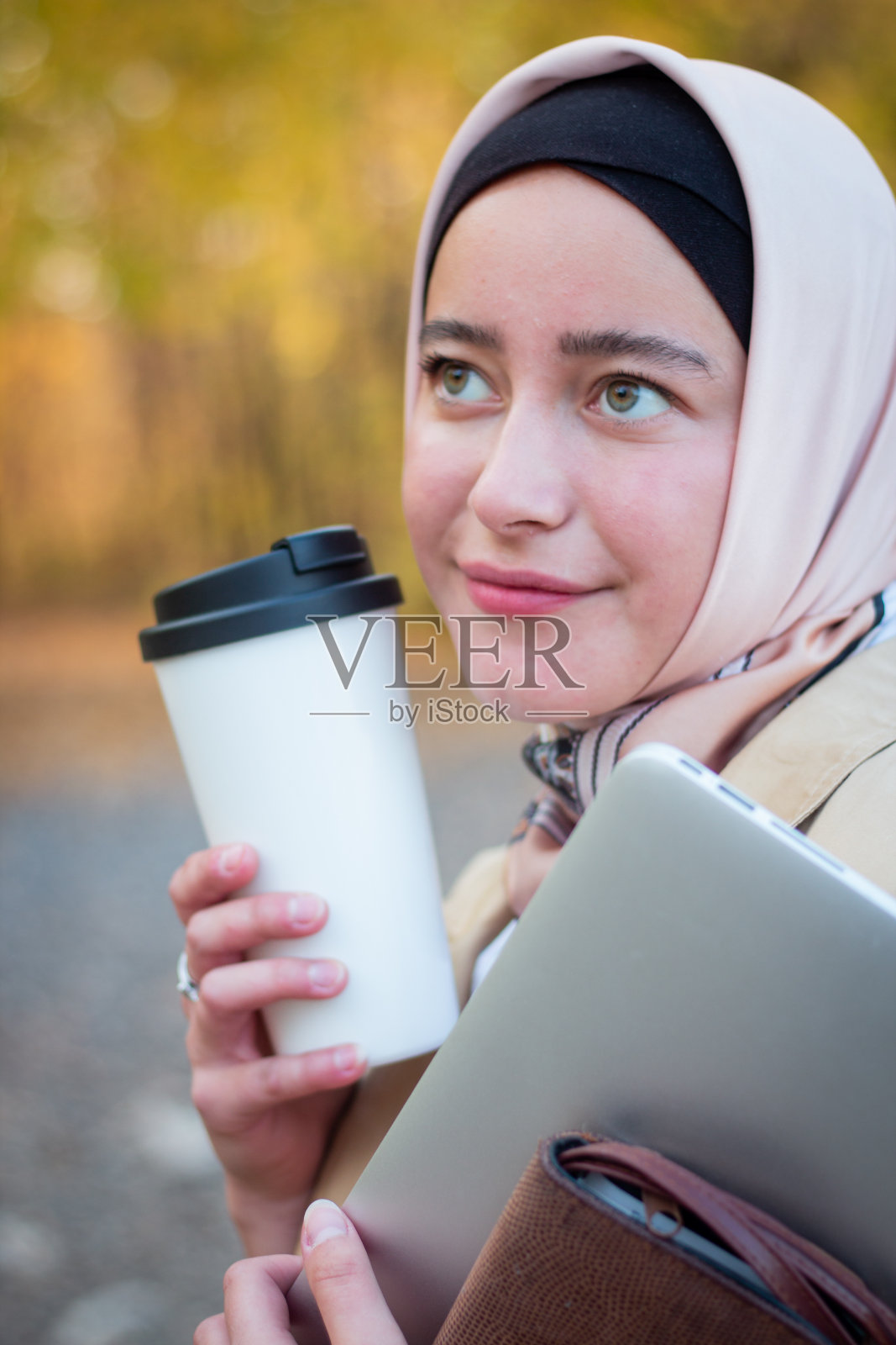 在公园里,一位戴着头巾、穿着米色风衣的穆斯林妇女拿着一杯咖啡和一台笔记本电脑站在那里。穆斯林商业女性。学生。关闭计划,脸。照片摄影图片