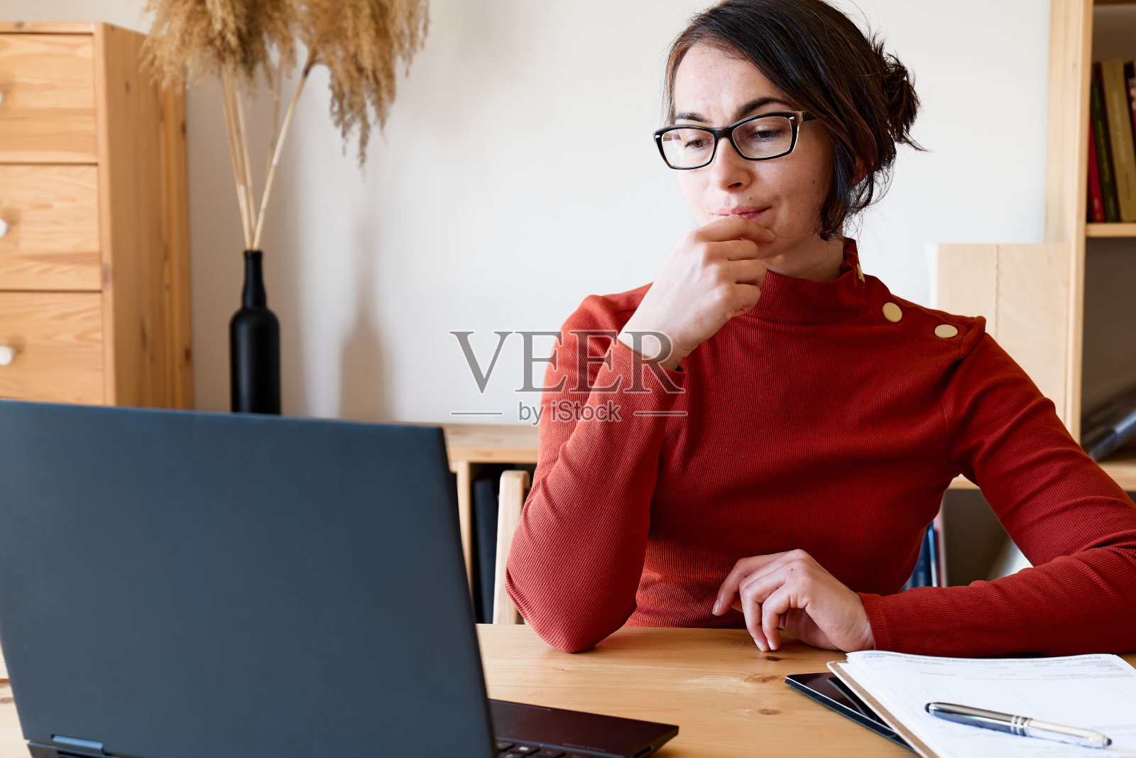 一位穿着红色高领毛衣、戴着眼镜的女士在窗边用笔记本电脑工作。远程工作的概念。照片摄影图片