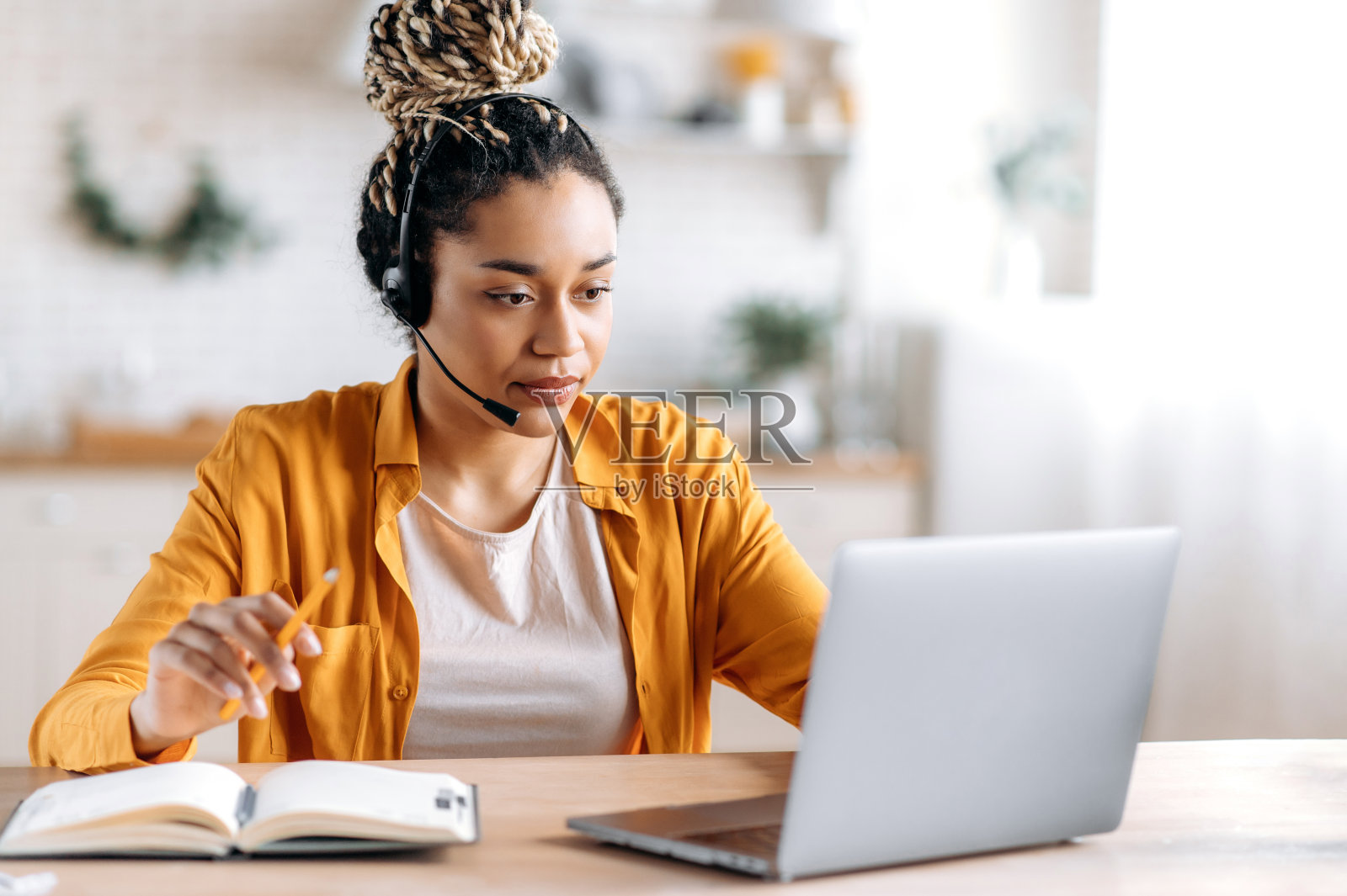 专注于聪明美丽的年轻非洲裔美国女人,梳着发辫,戴着耳机,穿着休闲服装,呼叫中心员工,自由职业者或经理,看着笔记本电脑屏幕,做笔记照片摄影图片
