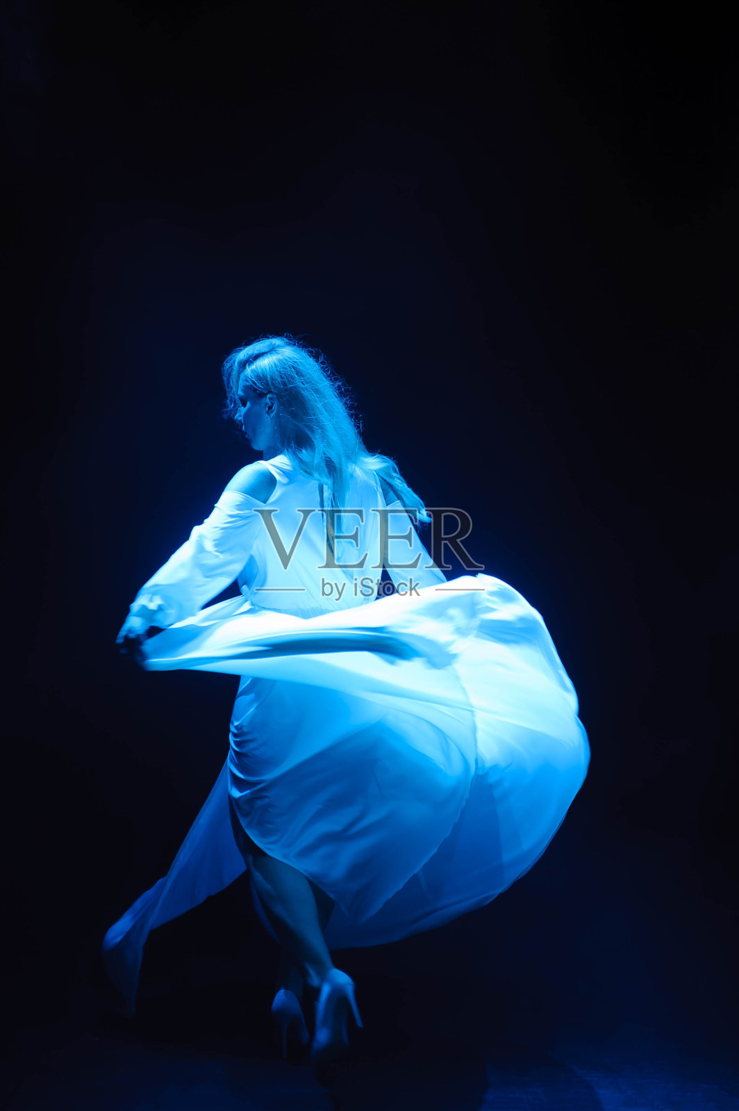美丽的女人在蓝色的灯光下穿着飘逸的白色裙子。流动的织物。轻盈的白布在风中飞舞。照片摄影图片