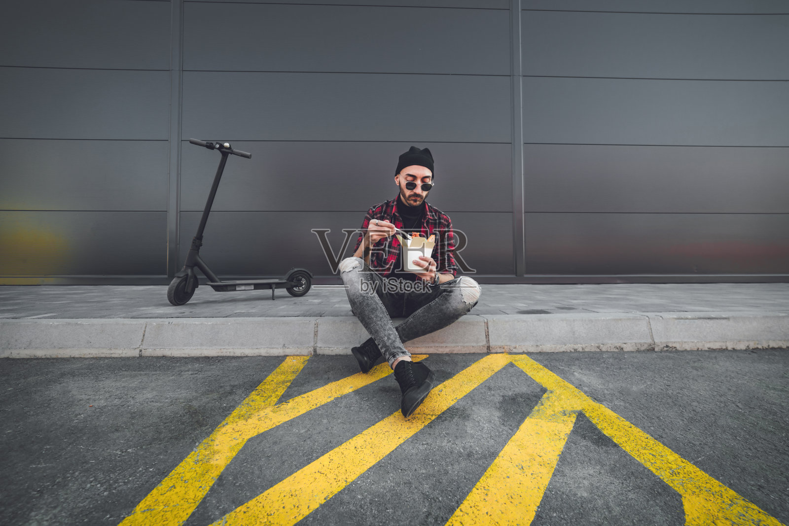 一个赶时髦的年轻人骑着电动滑板车坐在滑板车旁边休息,吃着美味的意大利面。他坐在人行道上,在商场的人行道上。照片摄影图片