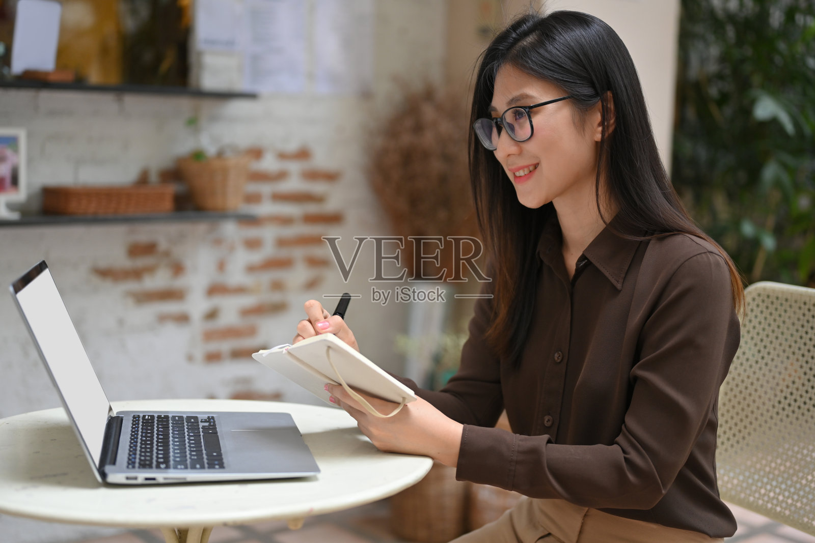 快乐的女员工一边在咖啡馆用笔记本电脑工作,一边在笔记本上写作照片摄影图片