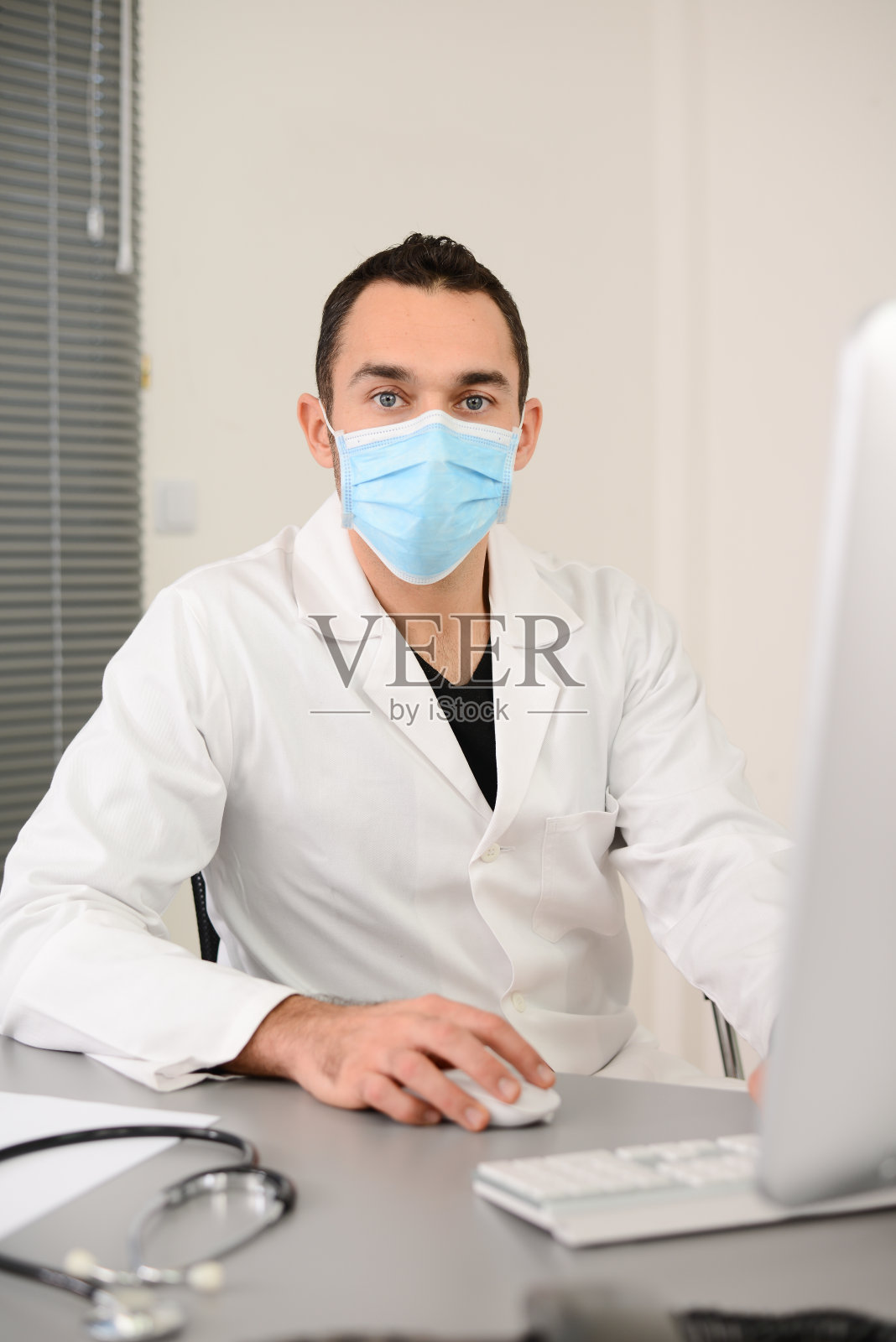 一个英俊的男人男医生在一个医疗实践办公室与外科口罩在电脑上工作的肖像照片摄影图片