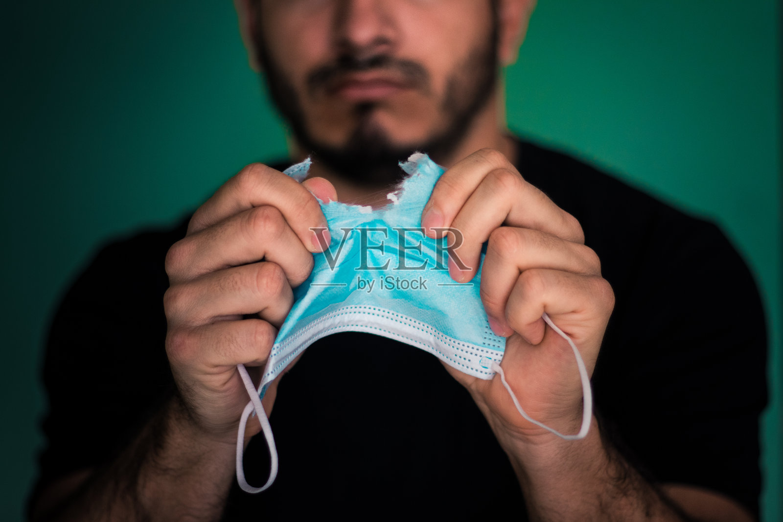 一名身穿黑色t恤的年轻成年男子撕扯着深蓝色背景的浅蓝色一次性口罩,口罩上有白色褶皱和条纹,暗指新冠肺炎大流行的主题照片摄影图片