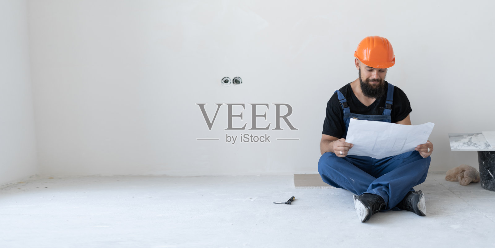 一个野蛮的男建筑工人正坐在地板上。穿着蓝色制服,戴着橙色面具。他手里拿着一张破旧的纸,上面有画。照片摄影图片