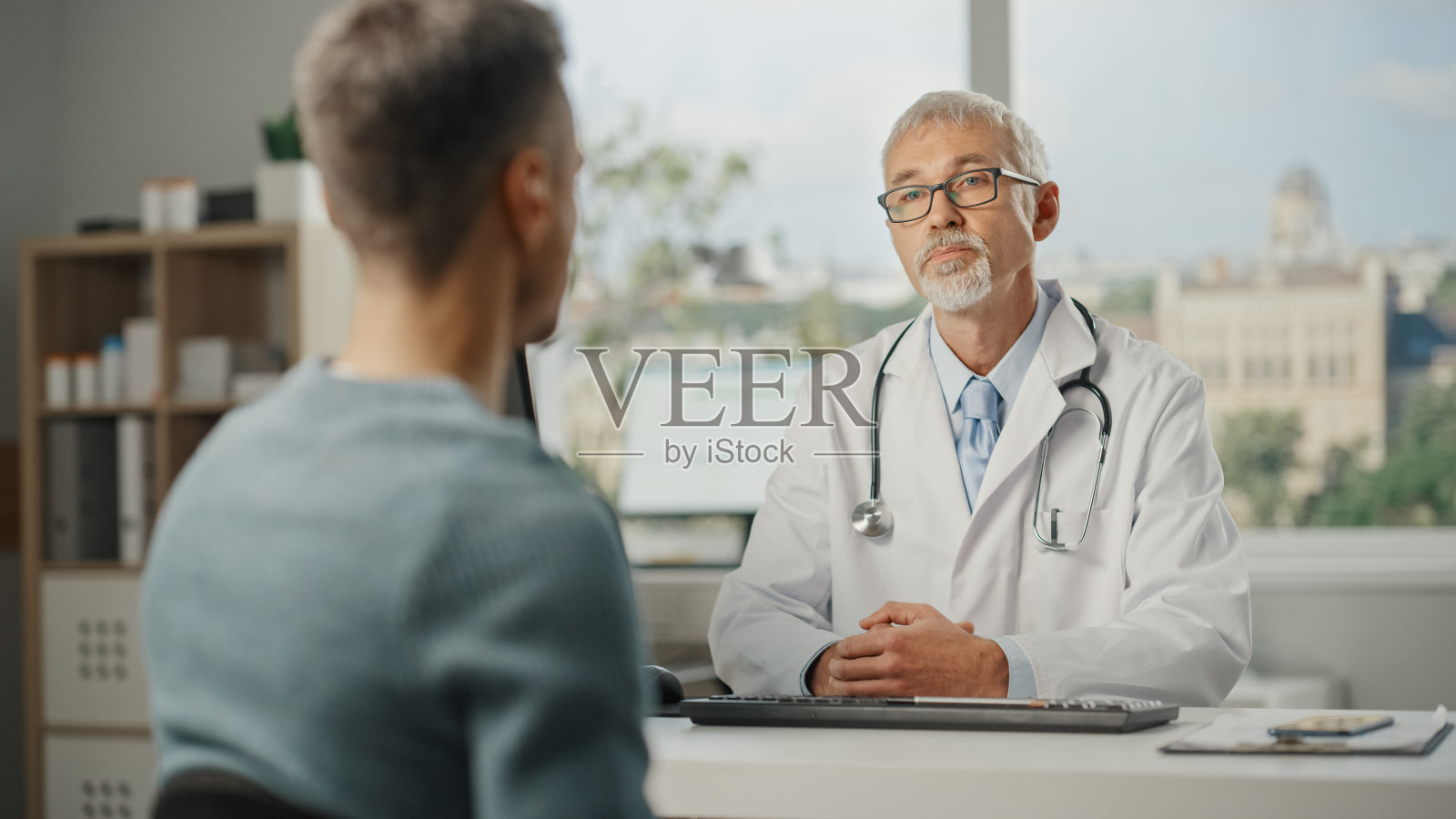 一位中年家庭医生正在一家健康门诊与年轻男性病人交谈。穿着白大褂的高级医生坐在医院办公室的电脑桌前。照片摄影图片