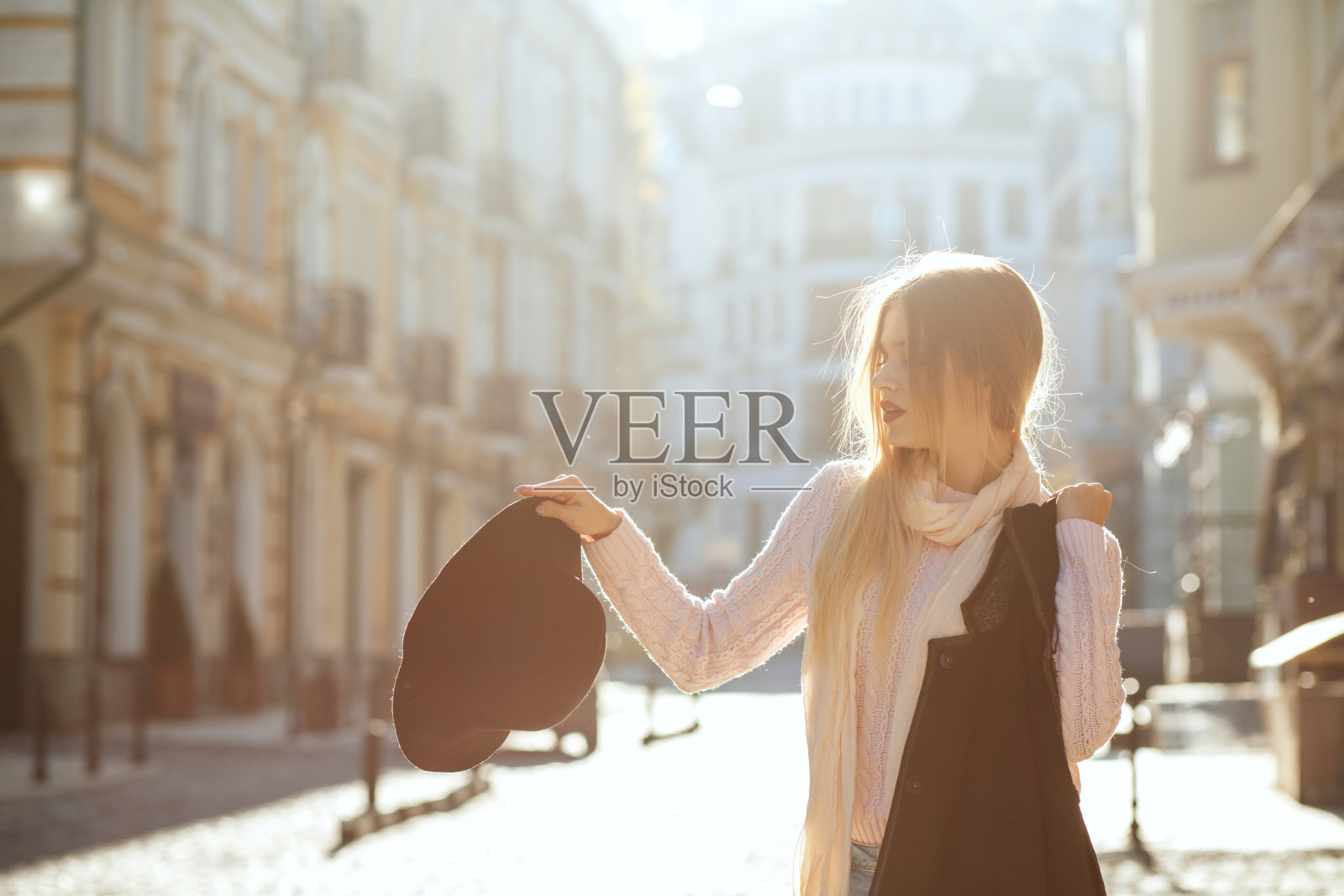 街头时尚概念:迷人的金发美女戴着帽子和外套在阳光老街摆姿势。空的空间照片摄影图片