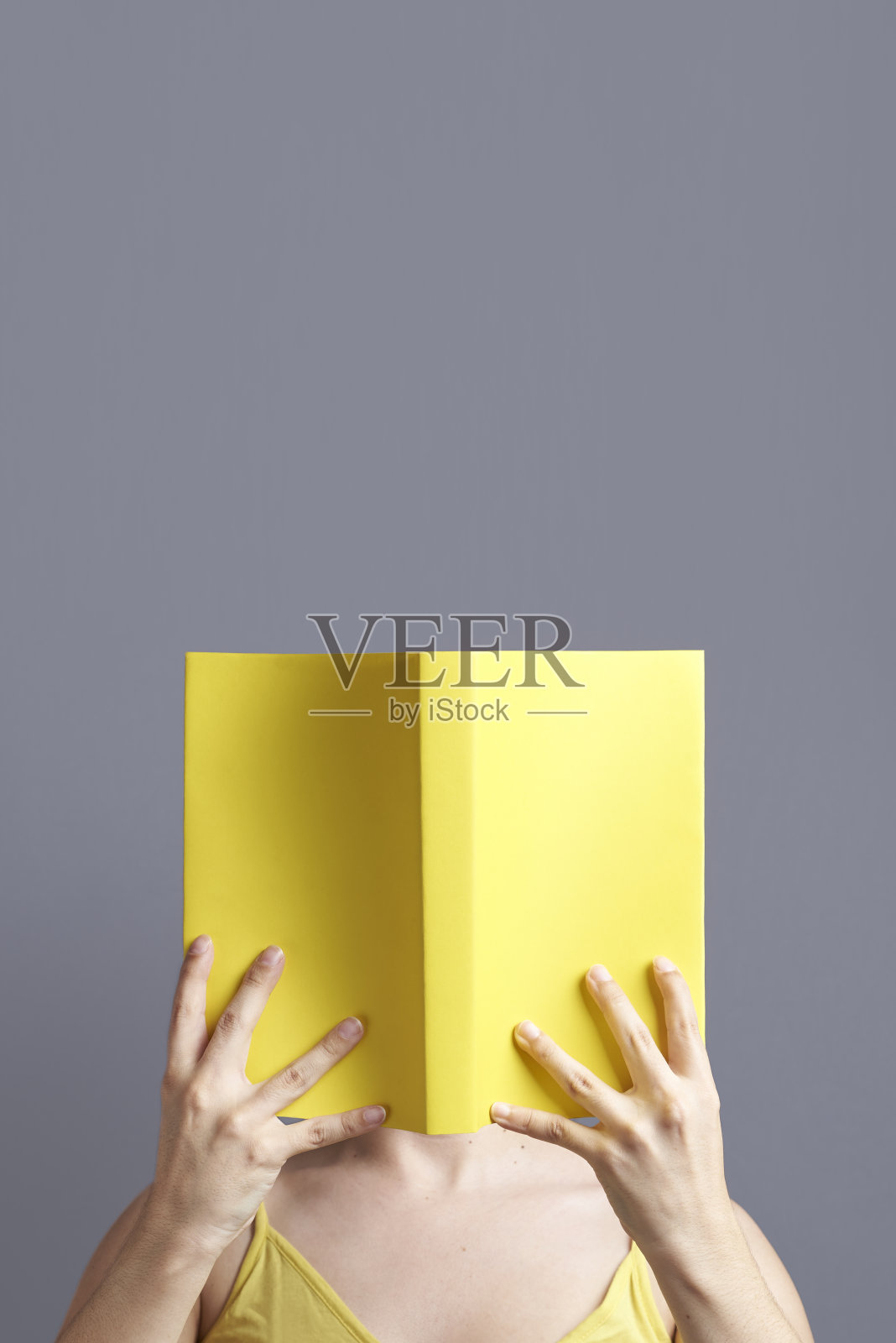 一个年轻的女人,她的脸隐藏在一本打开的黄色书后面,她正在阅读照片摄影图片