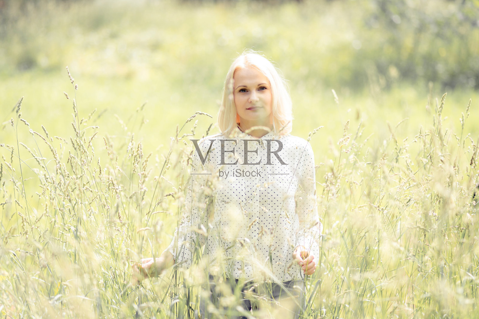 年轻漂亮的金发女人若有所思地看着相机在高草在阳光明媚的夏日。照片摄影图片