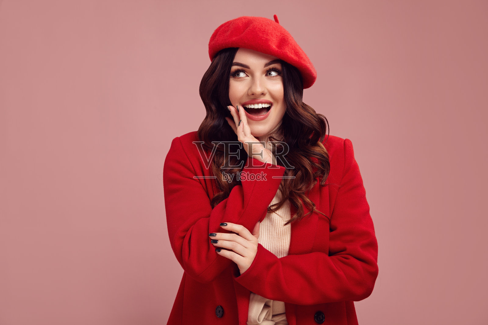 漂亮的白人惊讶的女人穿着红色的外套和贝雷帽表情惊讶地看着一边,嘴大张在惊讶,把她的手放在脸颊上孤立地站在粉红色的背景。照片摄影图片