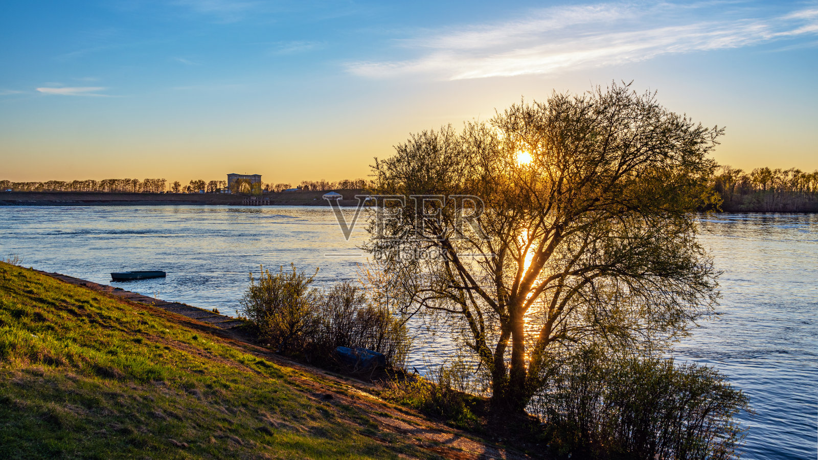河上的日落。水闸建筑的剪影在地平线上,一艘渔船的剪影和太阳耀斑通过一棵树在岸边。照片摄影图片
