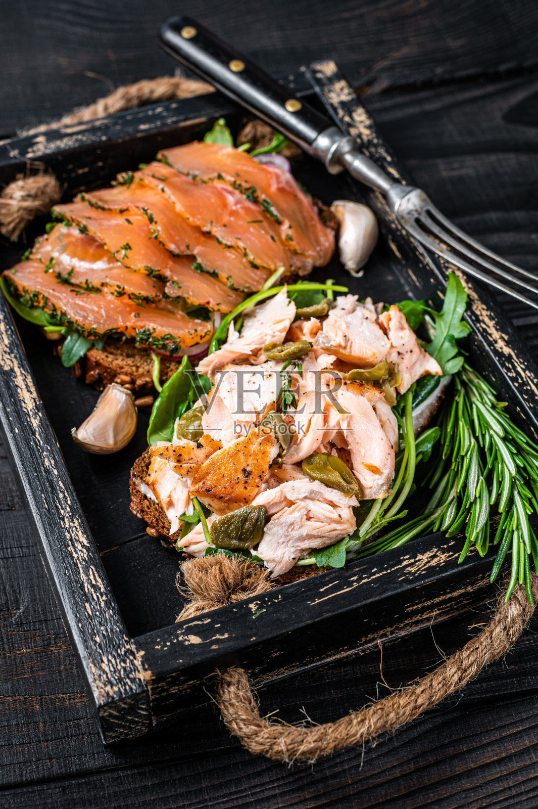 烤面包与冷热烟熏鲑鱼,芝麻菜在木托盘与香草。黑色木制背景。俯视图照片摄影图片