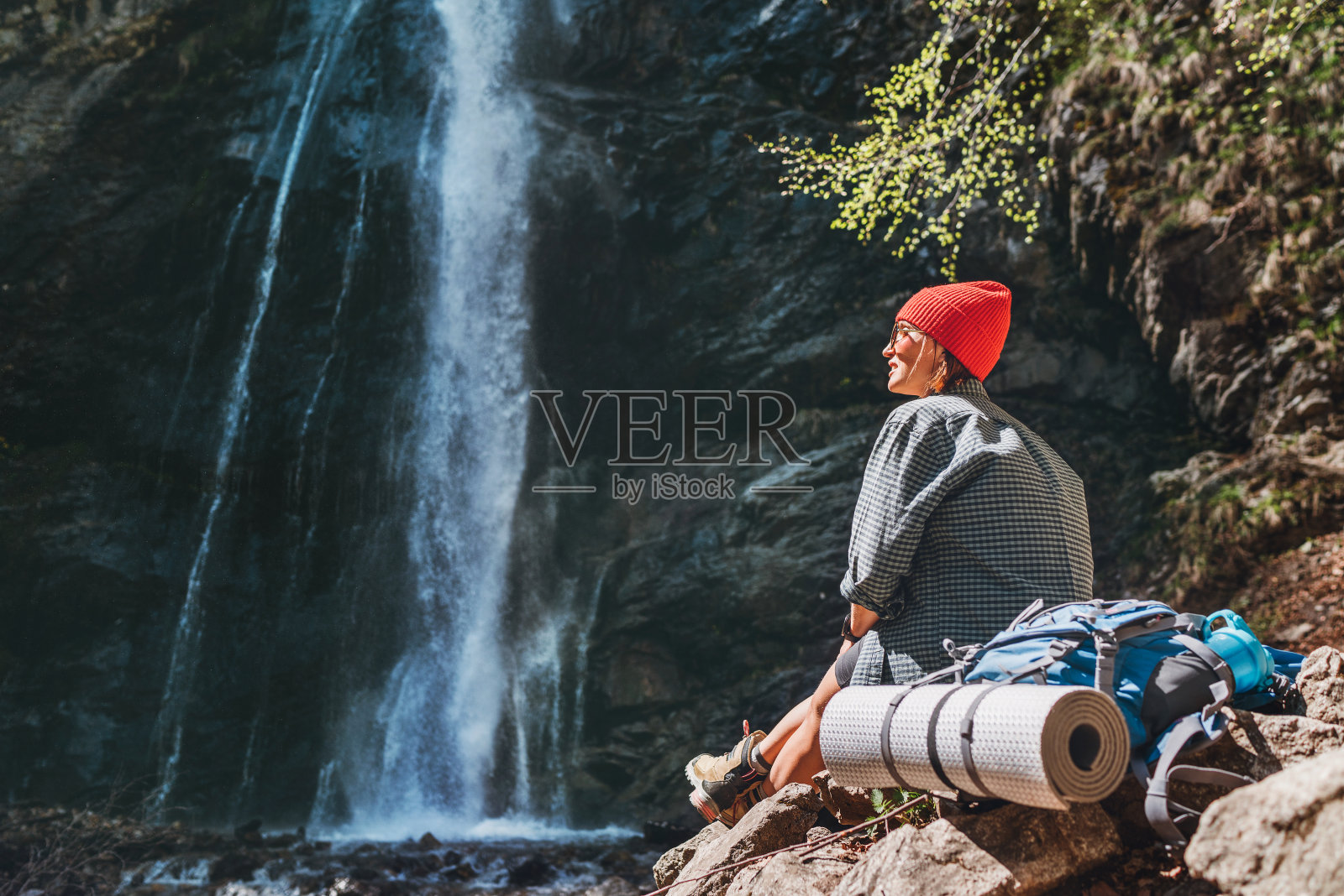 微笑的女人背包穿着活跃的徒步衣服坐在附近的山区河流瀑布,享受飞溅的自然力量。旅游、徒步和自然概念形象。照片摄影图片