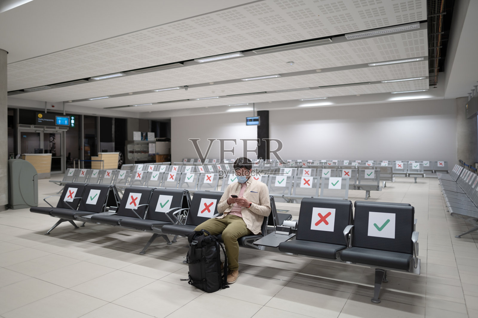 在塞浦路斯帕福斯国际机场,旅客在COVID-19危机期间坐在候机楼的座位上,戴着口罩,佩戴社交距离标记。在等待登机时使用手机照片摄影图片