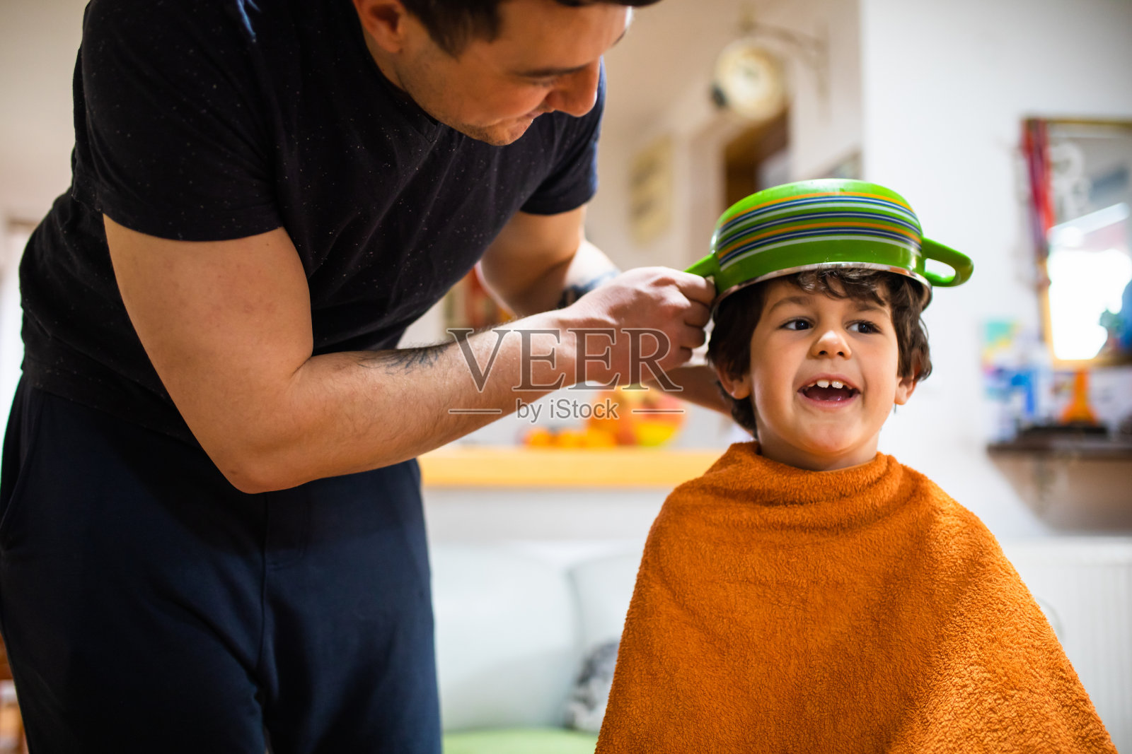 一位顽皮的父亲拿着一个罐子顶在儿子的头上,准备给他在家理发照片摄影图片