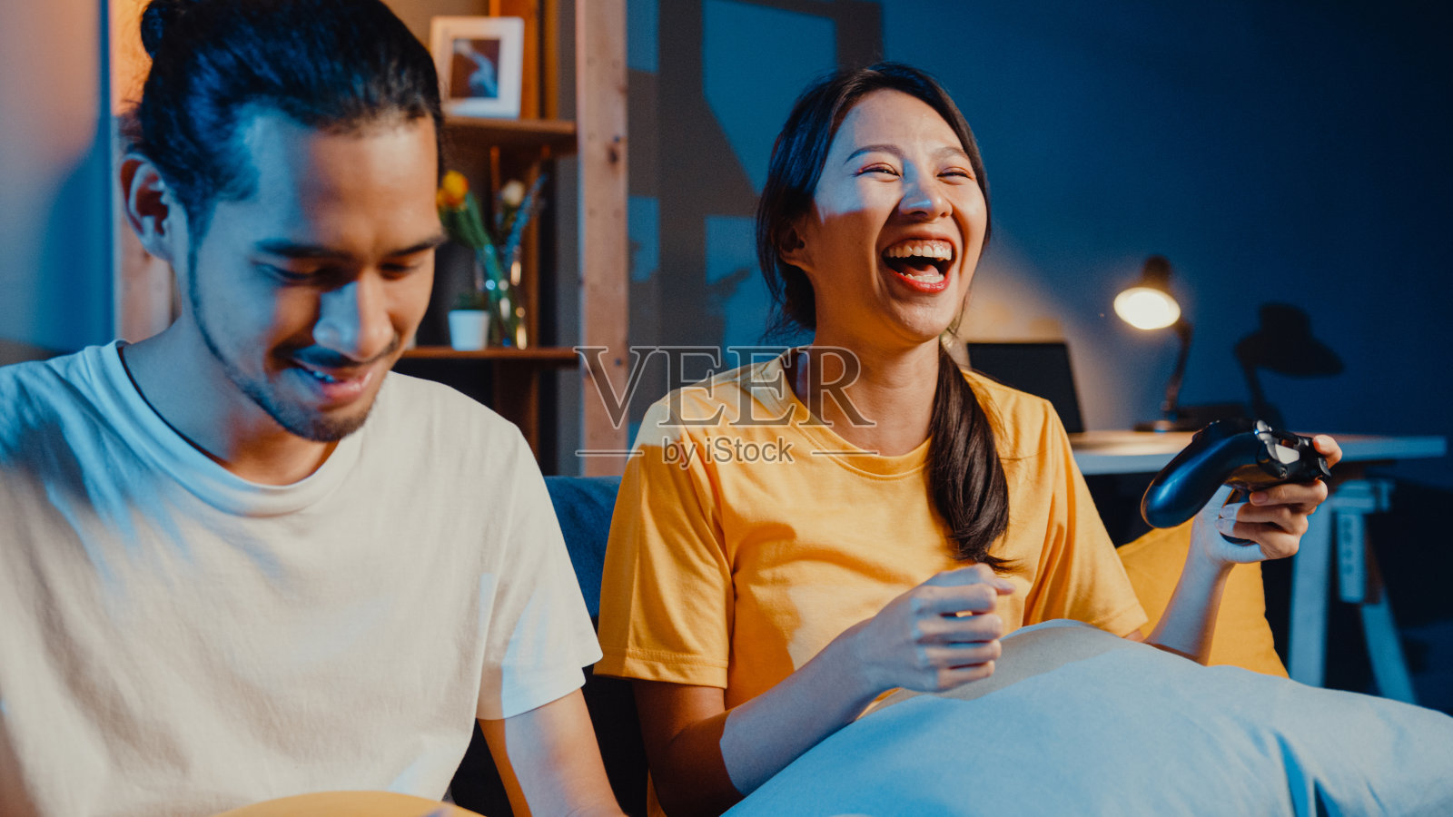 快乐亚洲年轻夫妇男女坐在沙发上使用操纵杆控制器玩电子游戏,晚上一起在客厅度过有趣的时光。照片摄影图片