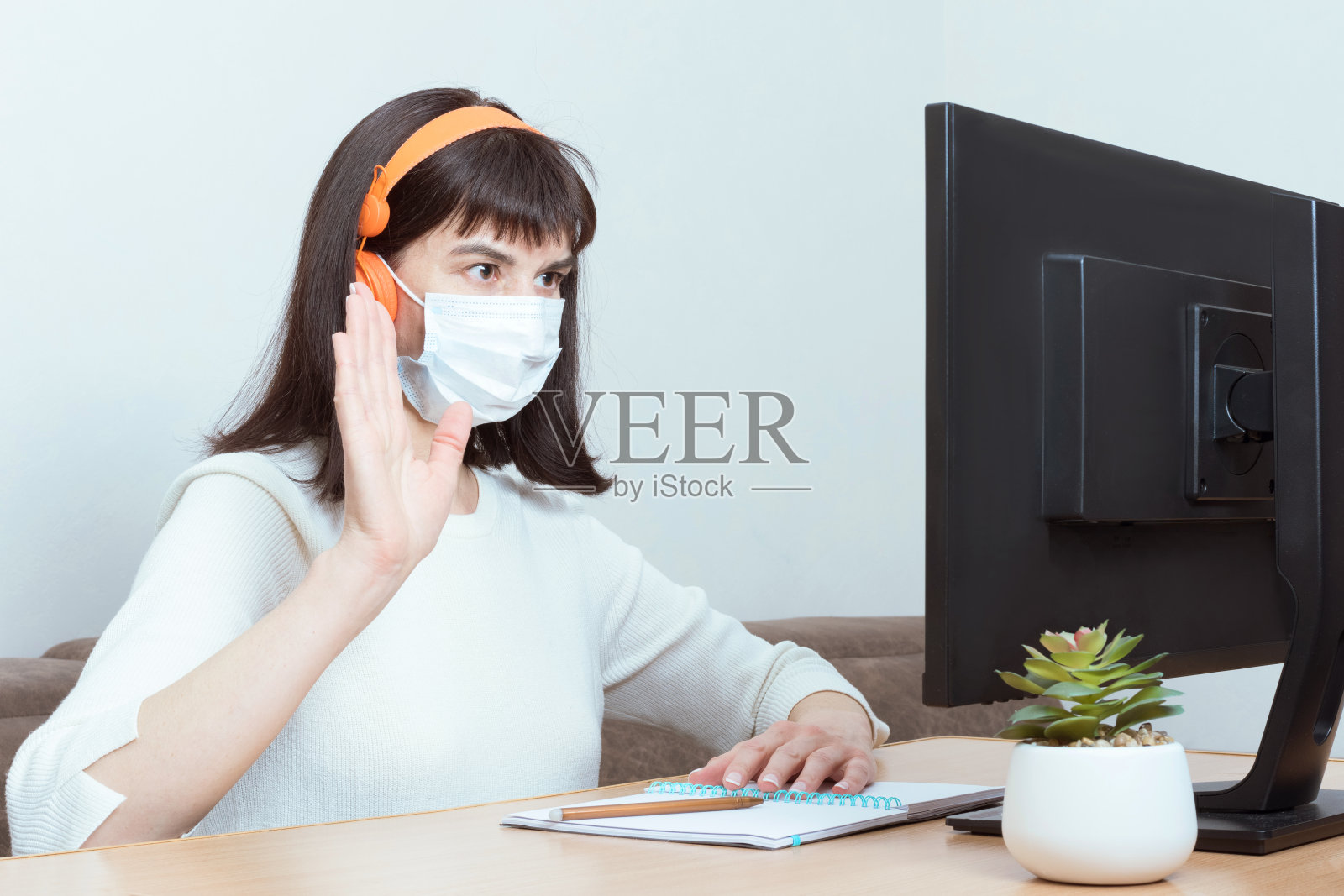 迷人的黑发女子戴着橙色耳机和医用口罩,视频通话和欢迎手势照片摄影图片
