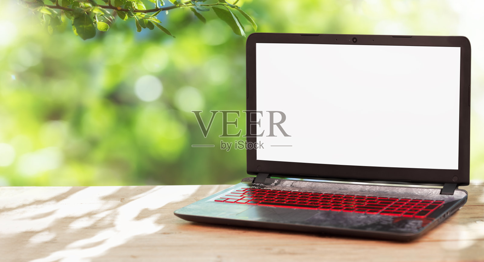 一台空白屏幕的笔记本电脑放在一张空木桌上,背景是稀疏的绿色繁茂的树叶。照片摄影图片