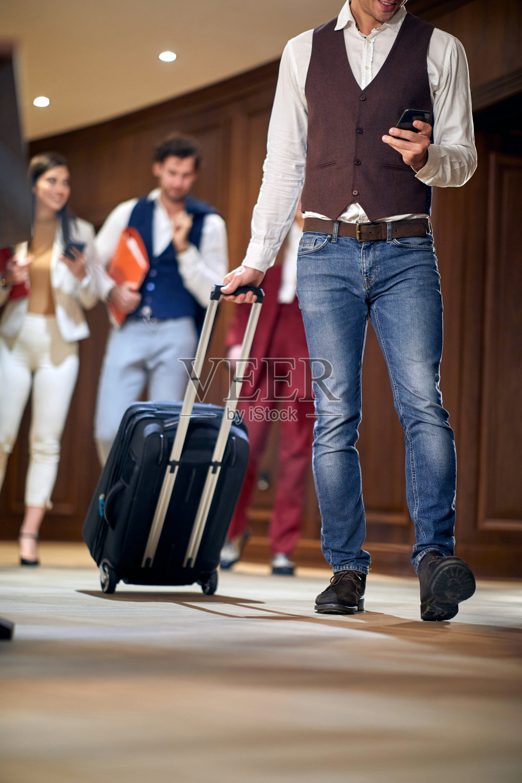 一群年轻人,商人,走过走廊。注意看一个拿着手机,带着轮式行李箱的男性。裁剪图像照片摄影图片