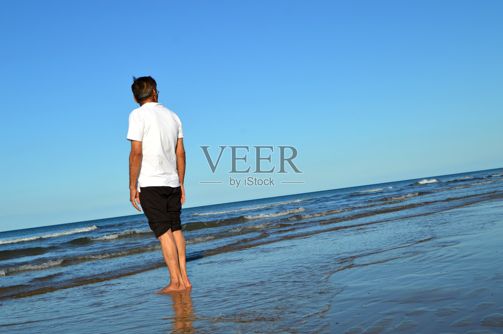 一位老人光着脚站在海滩的水里,背对着镜头照片摄影图片