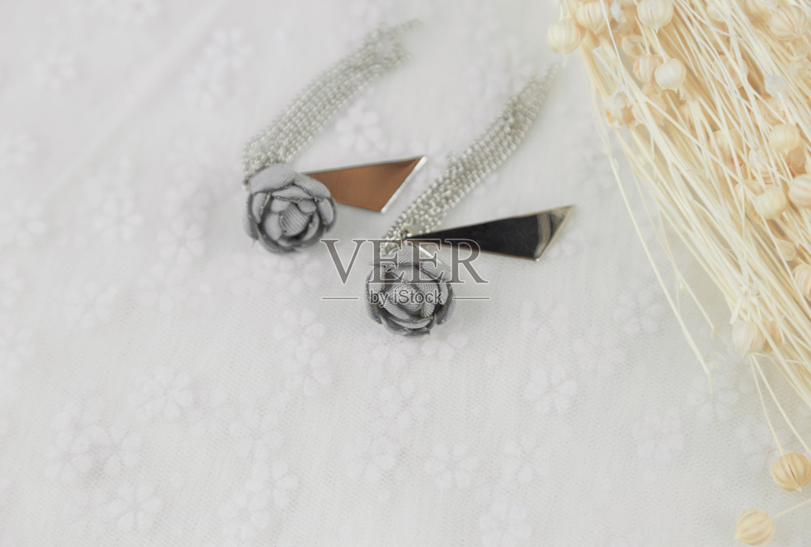 灰色吊坠耳环,它的形状是一朵花。在耳环旁边放一束花的标本。这些东西都放在薄纱上。这对耳环看起来非常优雅时尚。耳环以流苏和金属三角形装饰。照片摄影图片