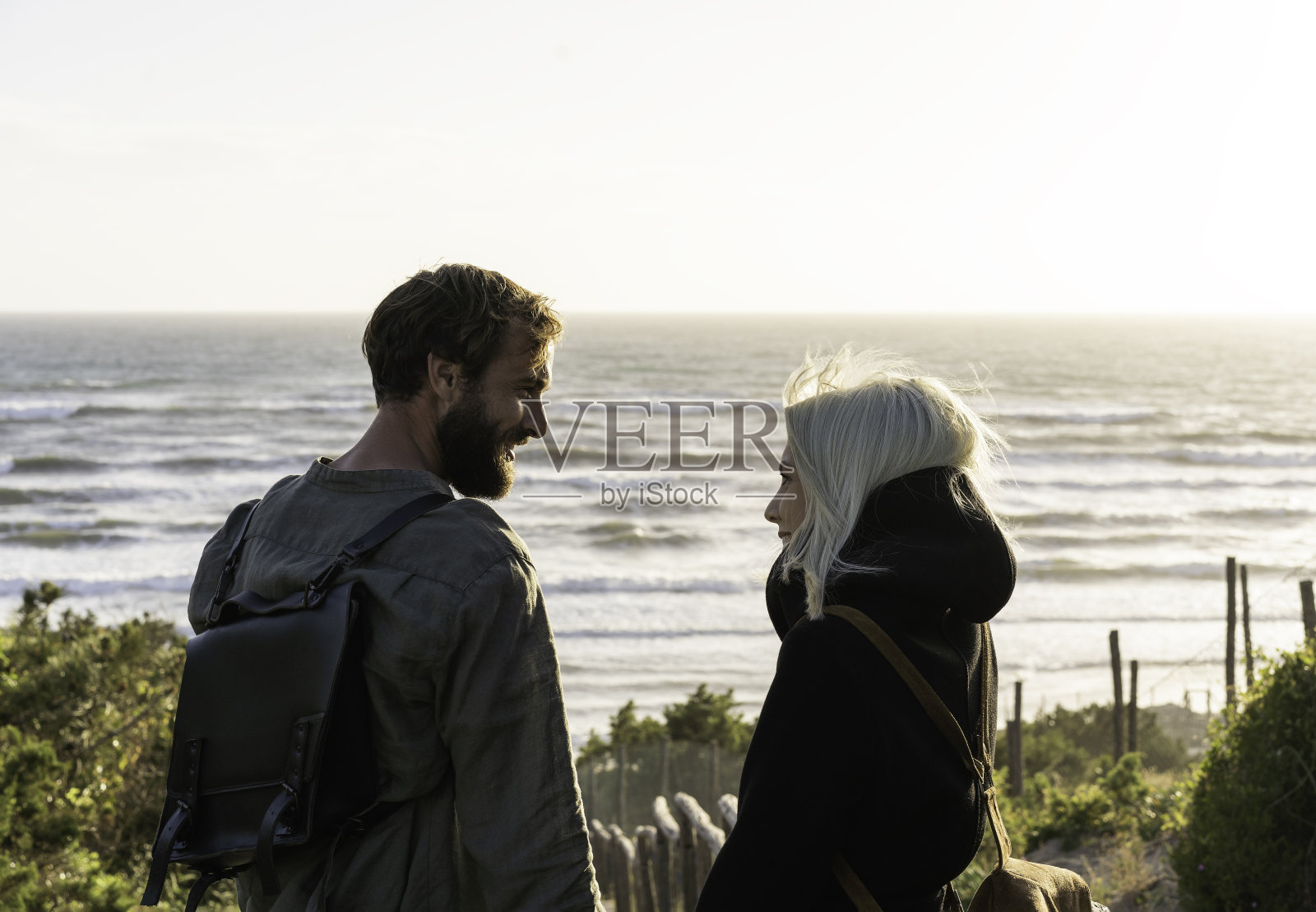 一对浪漫幸福的情侣站在沙滩上的木楼梯上,在日落时分,男朋友和女朋友背着复古背包看着对方的眼睛照片摄影图片