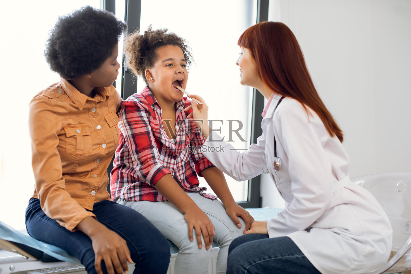 妈妈和女儿在医生办公室。白种人医生儿科医生,检查咽喉可爱的非洲少女,使用压舌板。说啊,喉咙痛,喉科概念。照片摄影图片