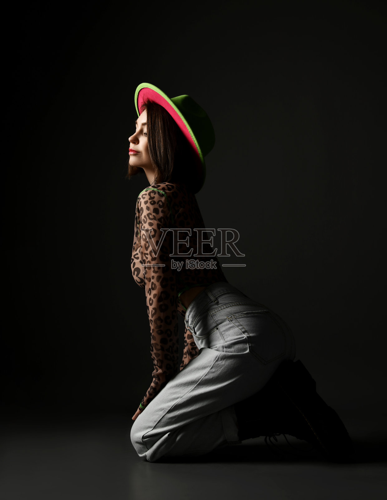 性感的年轻女人与大胸部穿着豹纹长袖,牛仔裤和帽子站在膝盖侧照片摄影图片