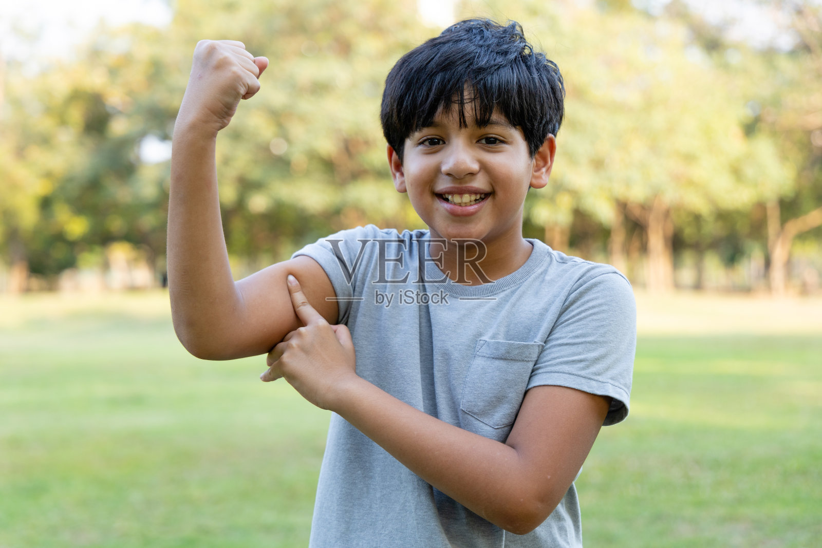 在公园里展示手臂肌肉的印度男孩。孩子微笑着与果酱做户外活动。健康的概念照片摄影图片