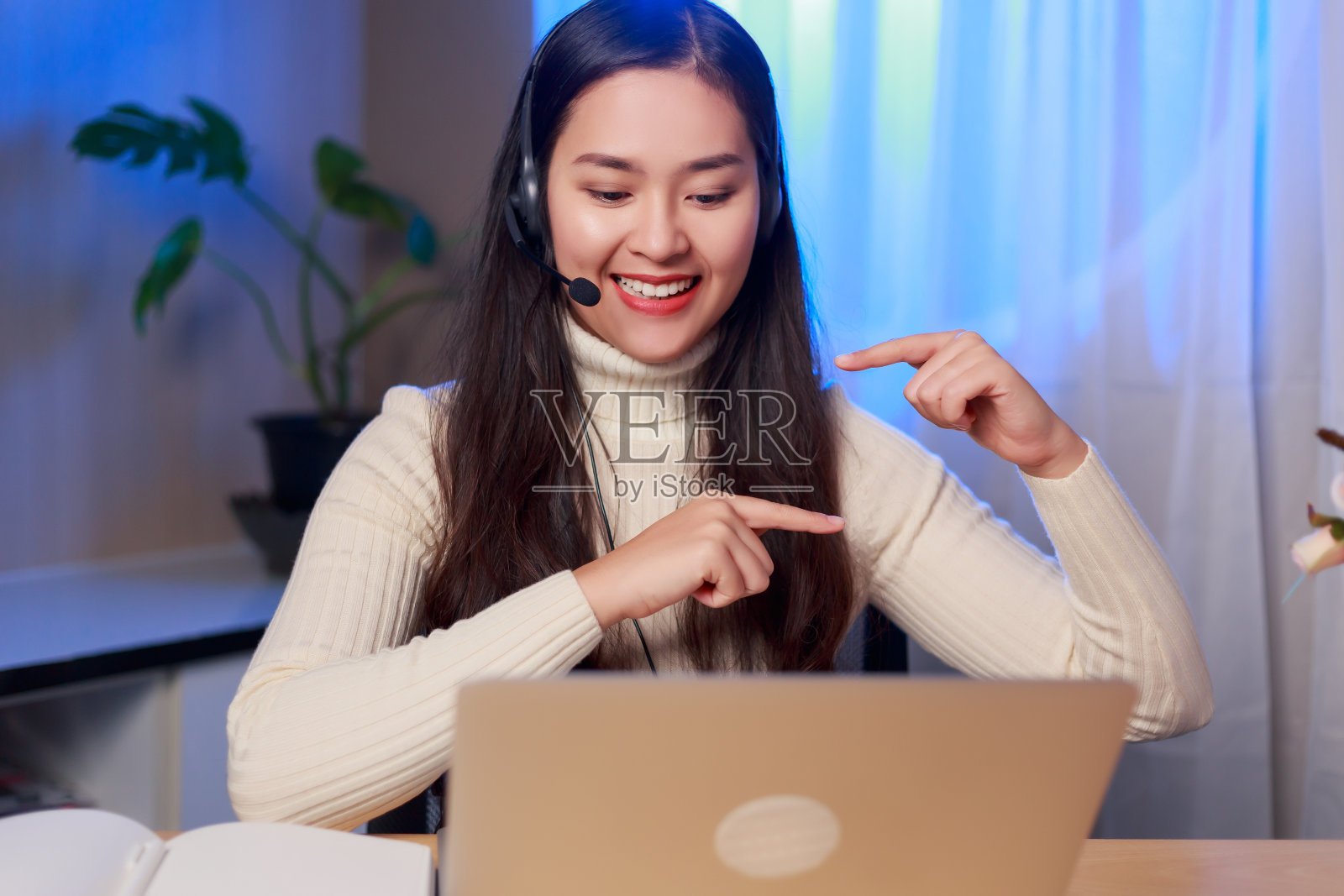 快乐年轻的亚洲女教练或操作员坐在笔记本电脑戴着耳机做在线工作在家里晚上照片摄影图片