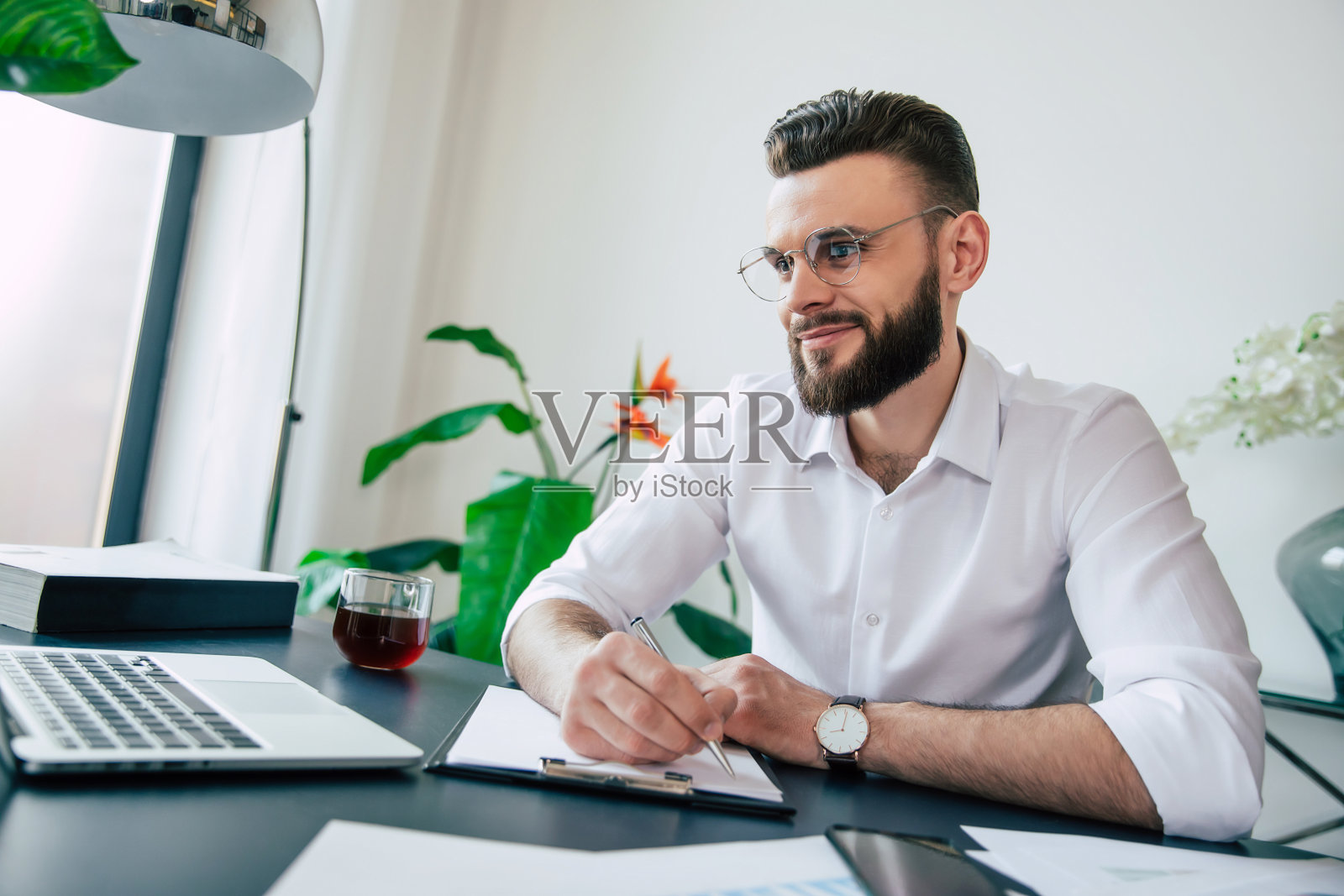 年轻、时髦、蓄着胡须、戴着眼镜、穿着白衬衫的商人正在用笔记本电脑处理办公室桌上的文件和文件。照片摄影图片