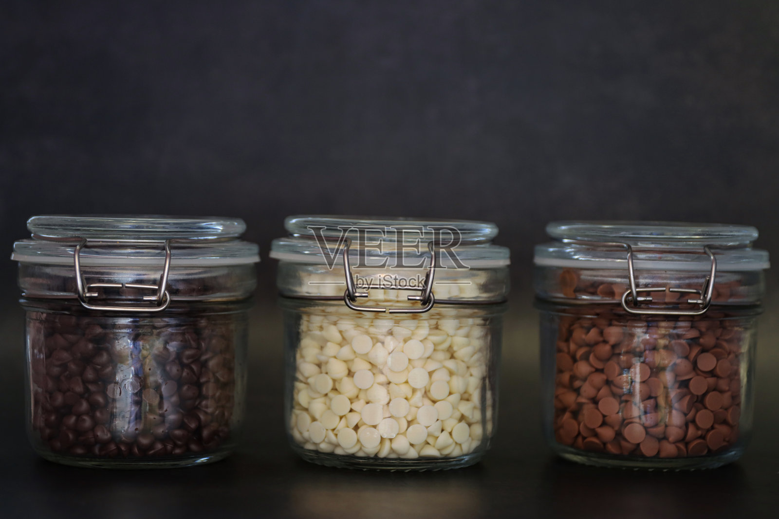 一排透明有盖的罐子,里面装满了牛奶、白巧克力和黑巧克力,背景是黑色的,聚焦前景照片摄影图片