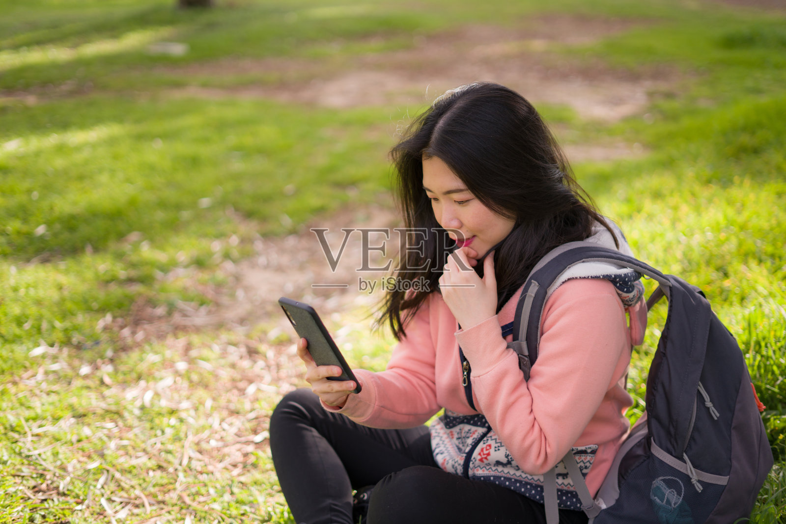 快乐的亚洲女人在城市公园使用手机-年轻甜美美丽的中国女孩坐在绿色草地上,用手机享受社交媒体照片摄影图片