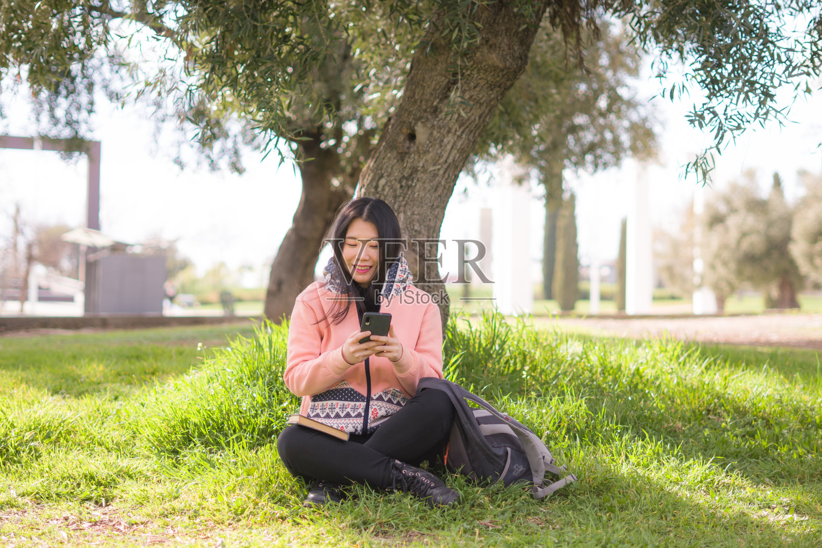 快乐的亚洲女人在城市公园使用手机-年轻甜美美丽的中国女孩坐在绿色草地上,用手机享受社交媒体照片摄影图片