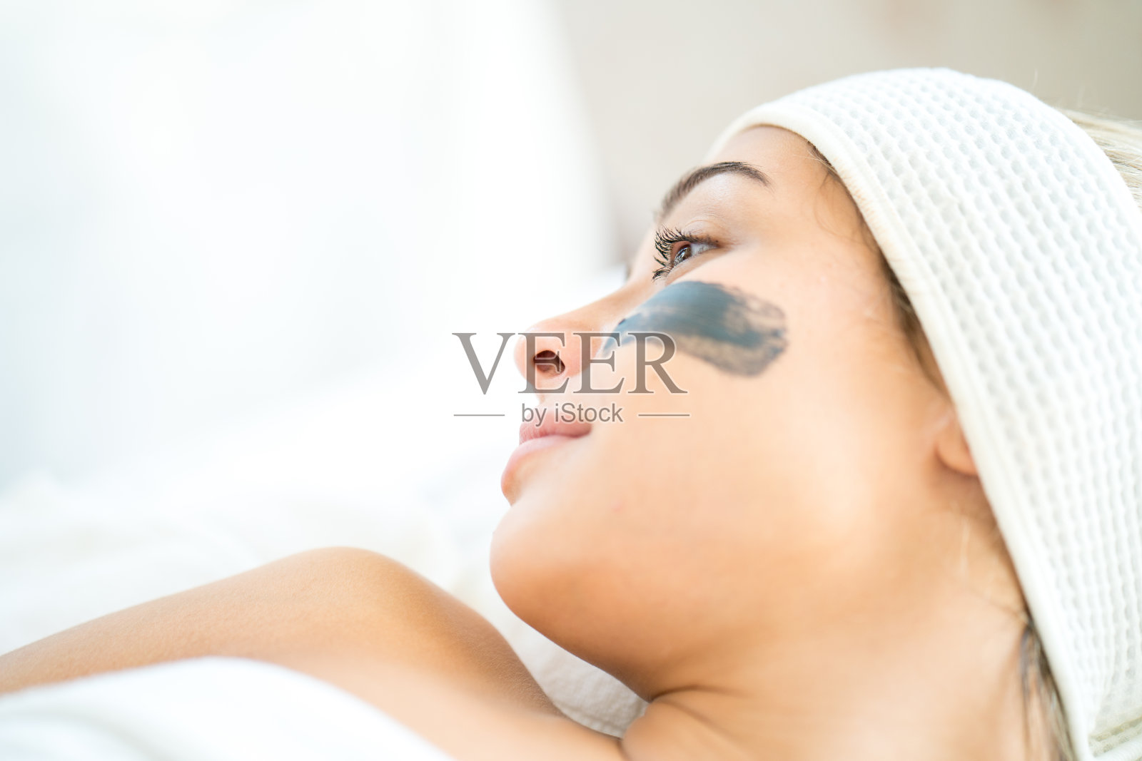 水疗中心的工作人员正在把护肤霜涂在脸上。一个年轻女子在水疗沙龙里用面具蒙面。一位年轻的白种女子在美容院按摩。照片摄影图片