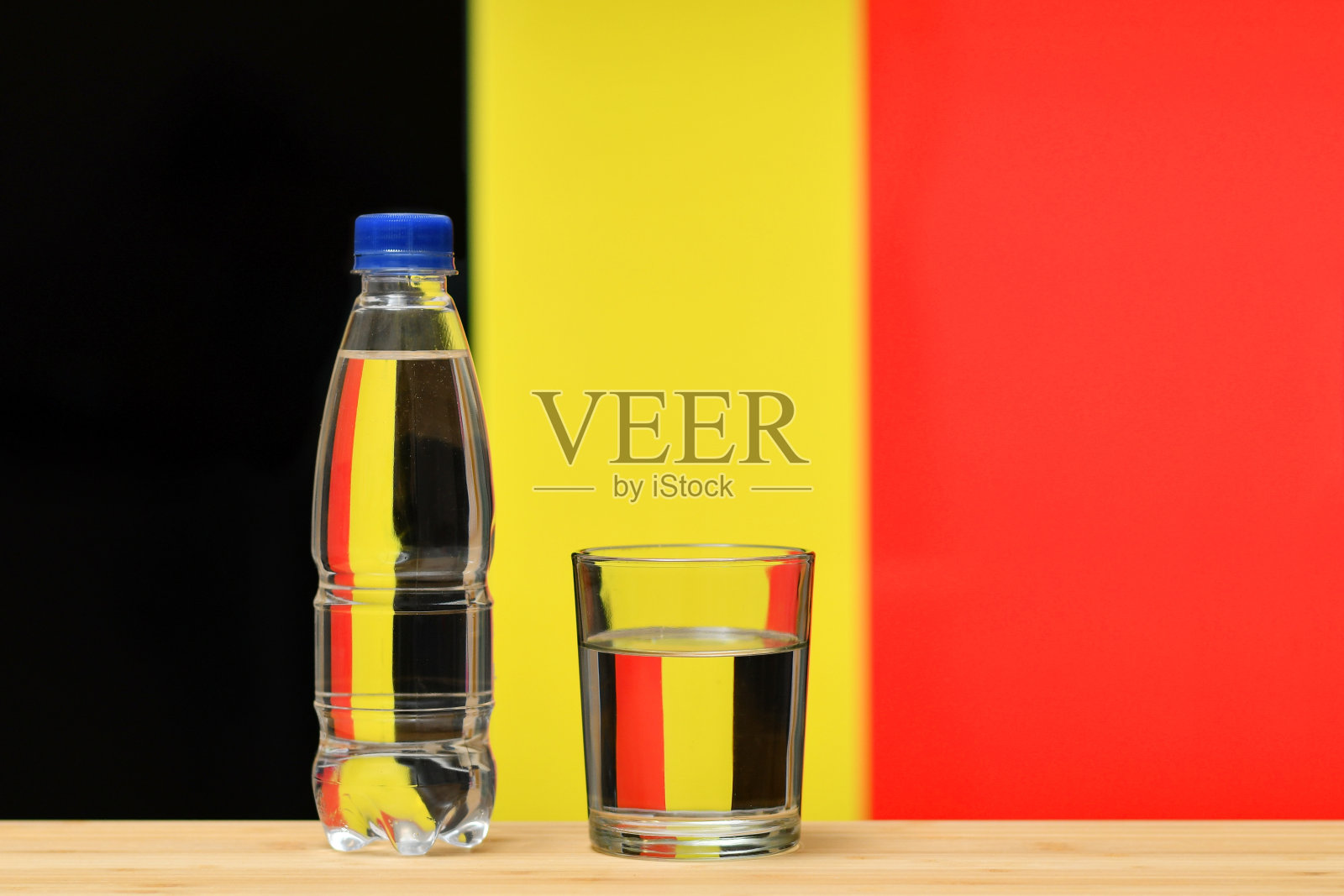 以比利时国旗为背景的桌子上放着一瓶干净的饮用水和一个玻璃杯。照片摄影图片