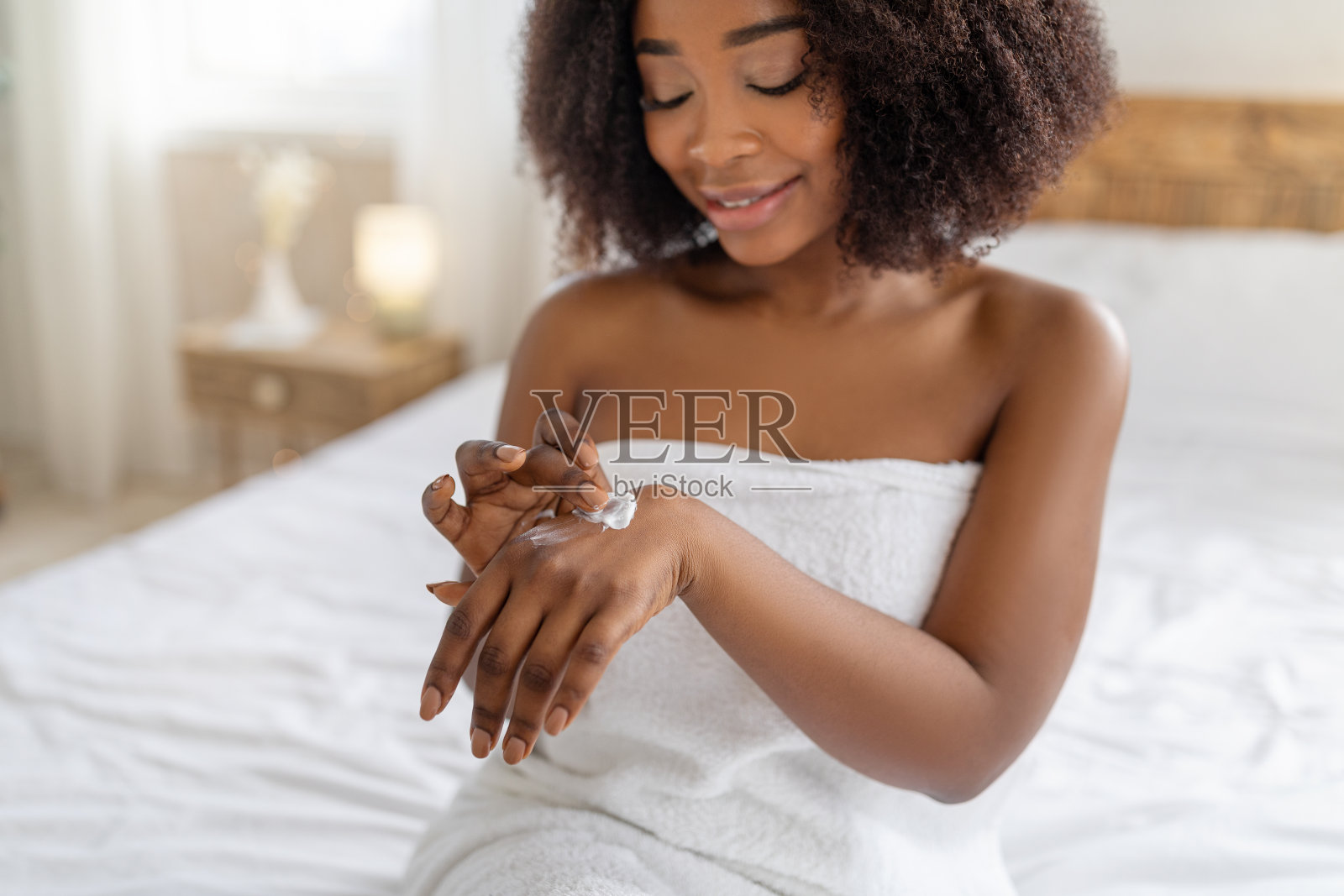 感性的非洲裔美国妇女坐在床上在浴巾,应用滋养霜在她的手在家里照片摄影图片