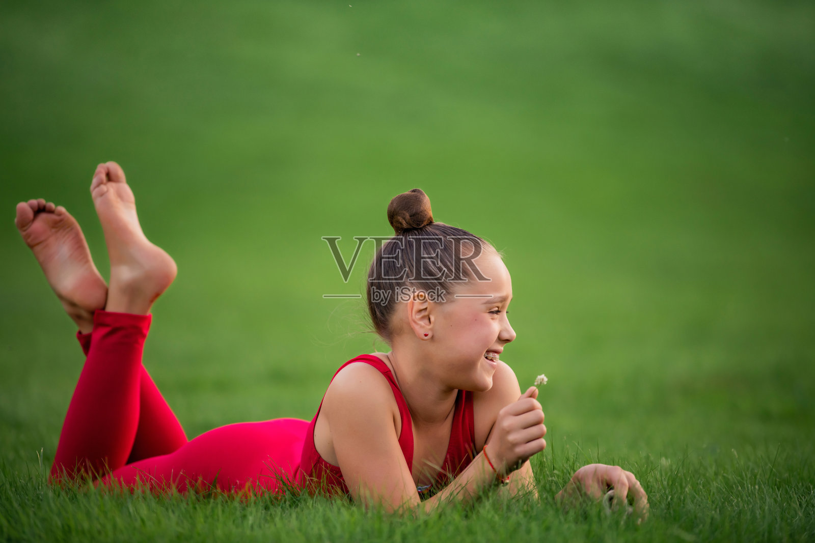 穿着鲜红色工作服的女学生体操运动员正在公园的草地上休息,手里拿着三叶草花,笑着。她戴着牙套。照片摄影图片