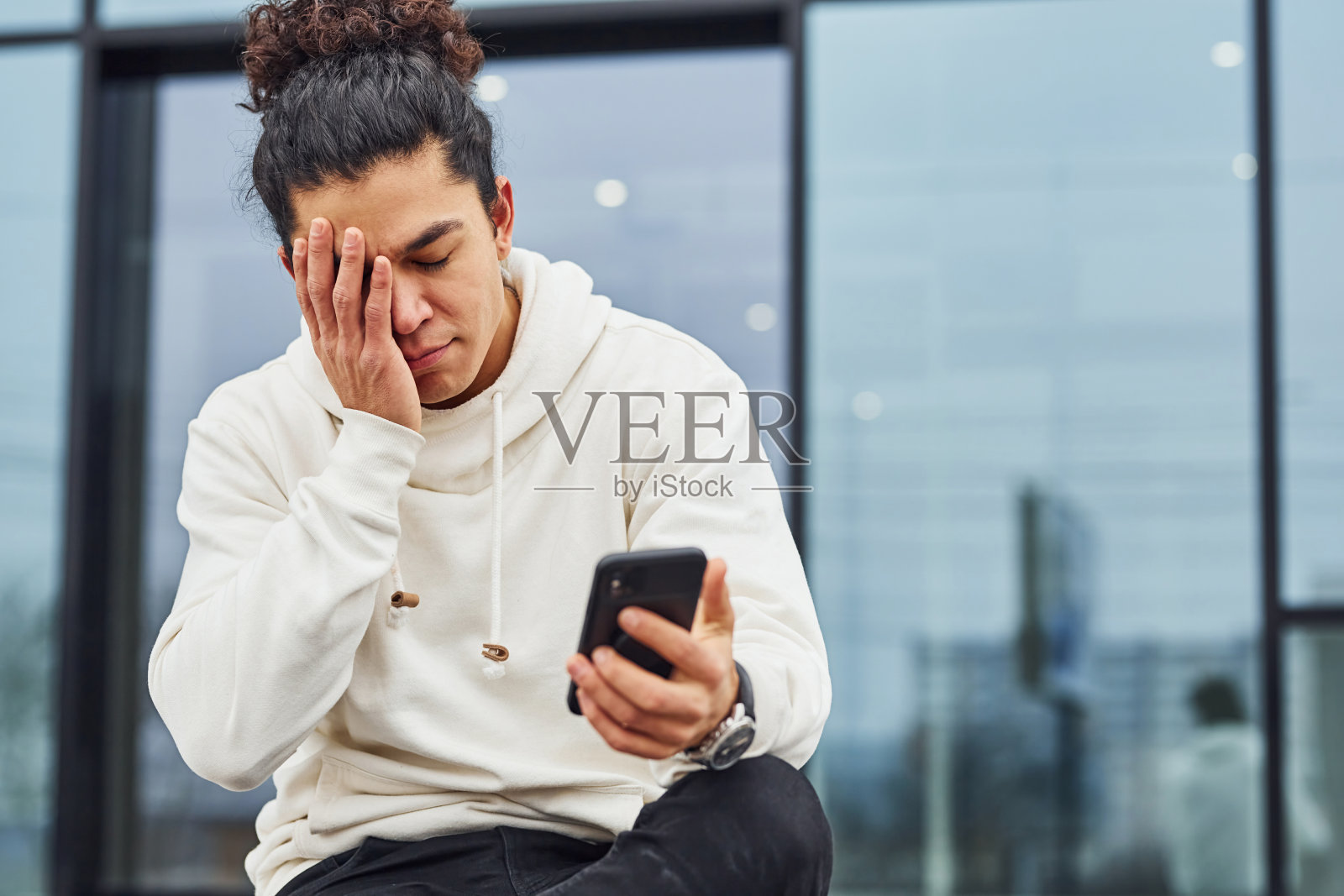 英俊的年轻人与卷曲的黑发坐在街上与智能手机对着大楼照片摄影图片