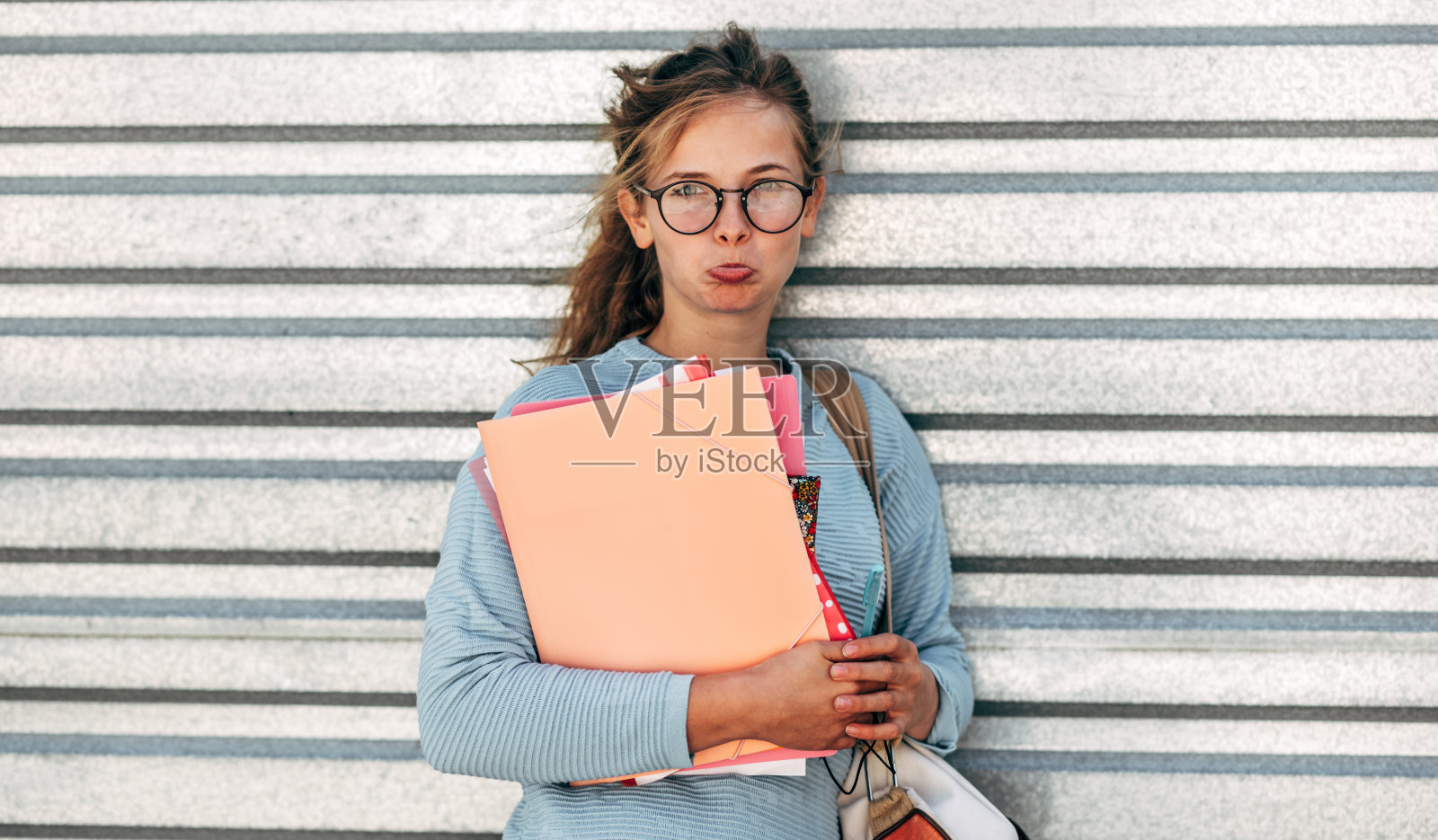 一个闷闷不乐的女学生在一天的大学生活后背着很多书的肖像。一个很伤心的年轻女人,穿着蓝色毛衣,戴着眼镜,拿着书和文件夹在街上摆姿势。照片摄影图片