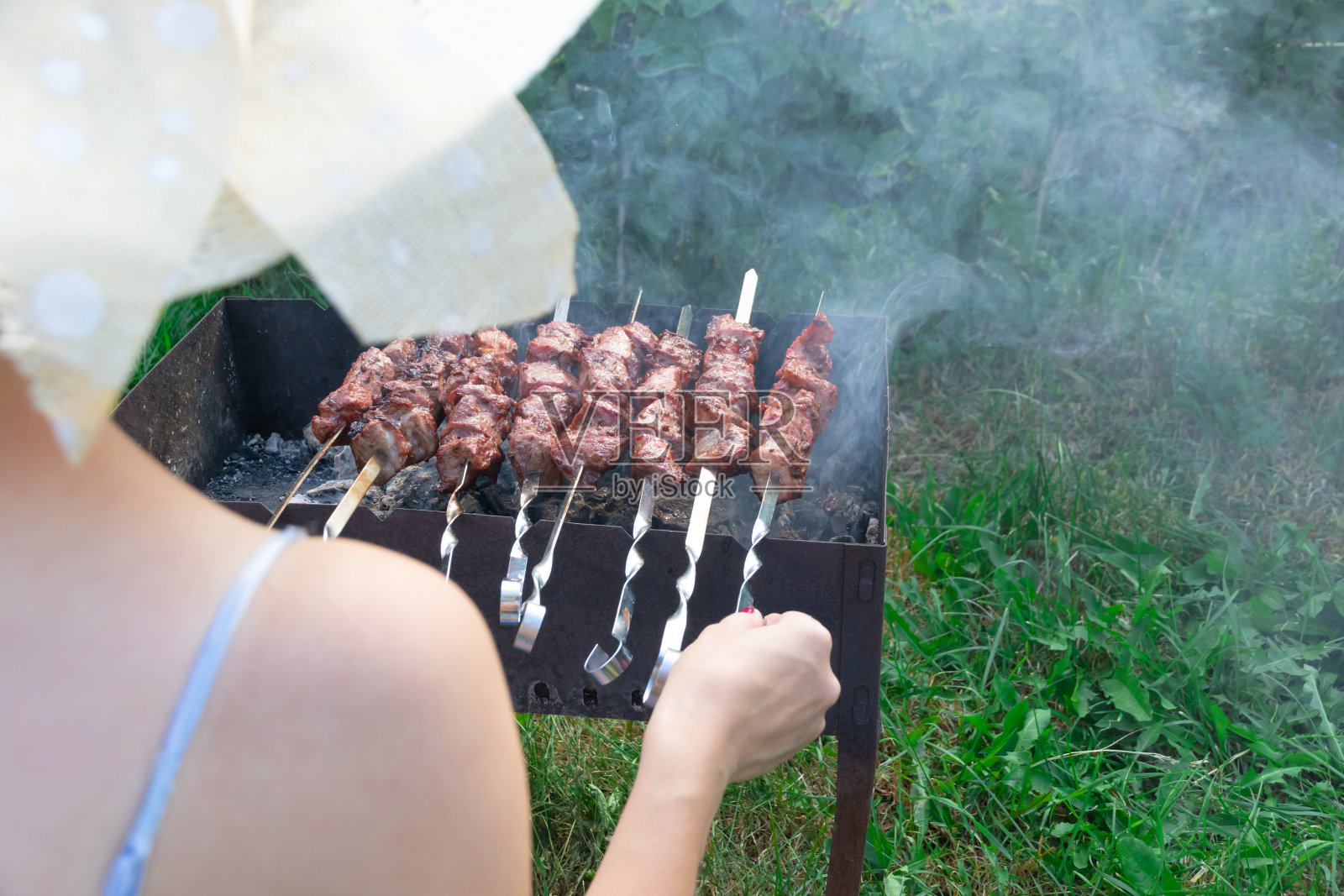 在一个阳光明媚的夏日,一个戴着草帽的女孩在绿色的树叶的背景下,在烤架上准备着芳香扑鼻的鲜肉照片摄影图片