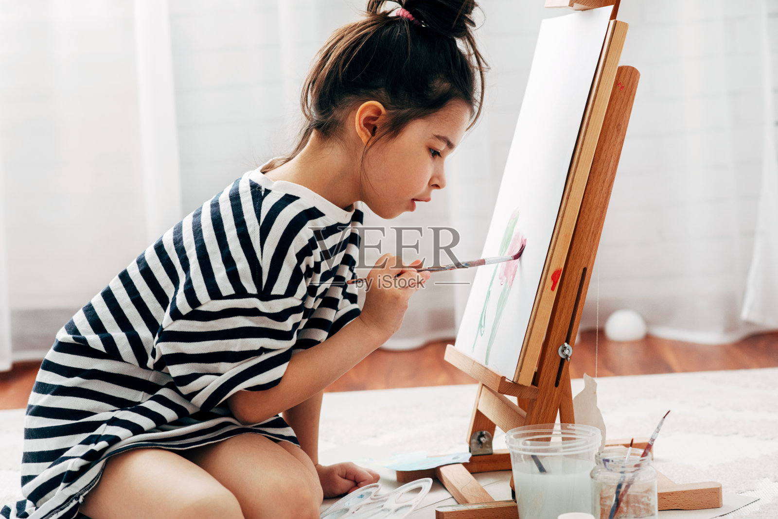 一个小女孩在家里的画架上用画笔在纸上作画的侧视图。一个可爱的孩子在禁闭期间坐在地板上在她房间里画画。照片摄影图片