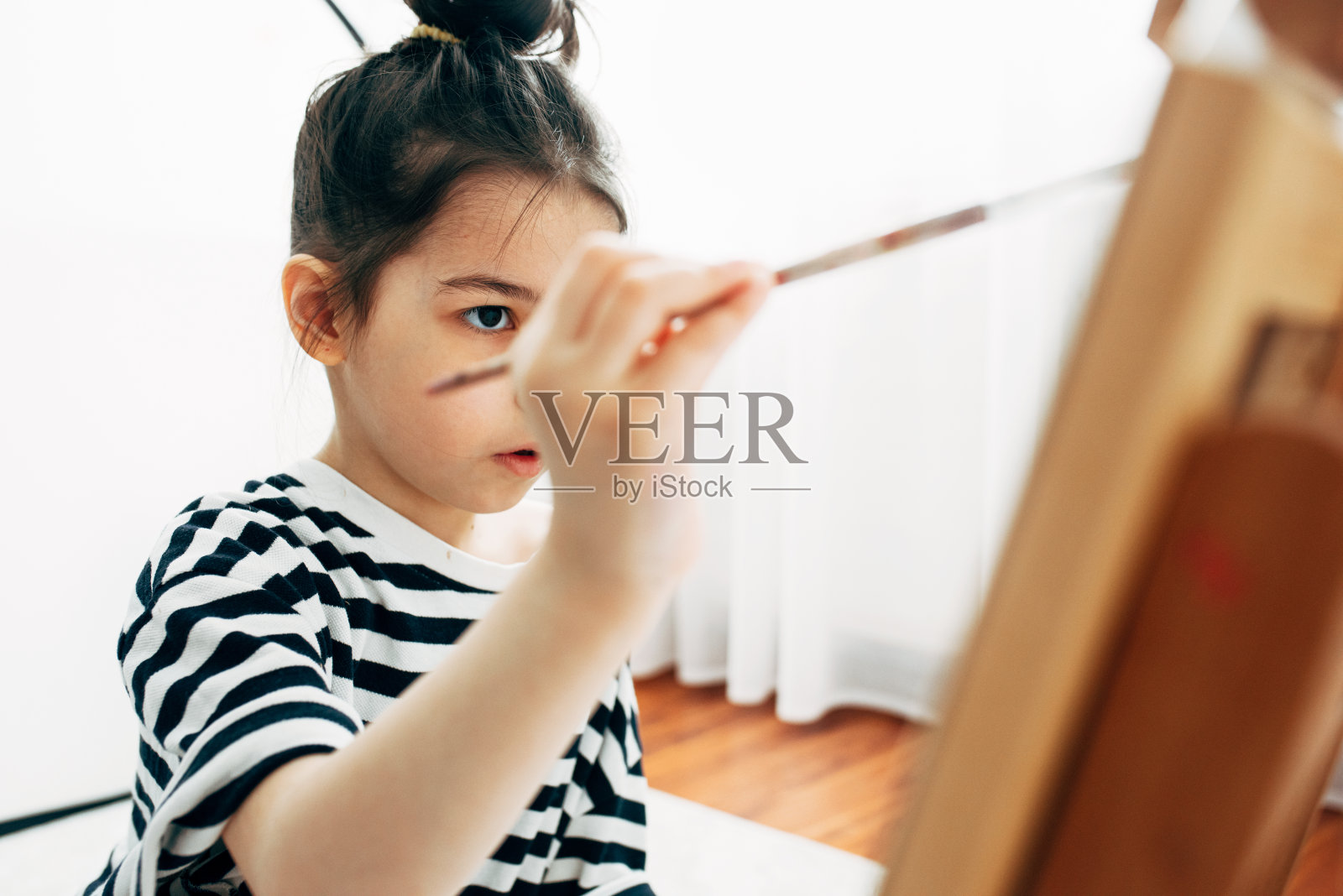 一个可爱的小女孩在家里的画架上用画笔在纸上作画的特写肖像。一个孩子坐在地板上,在她的房间里画画。照片摄影图片