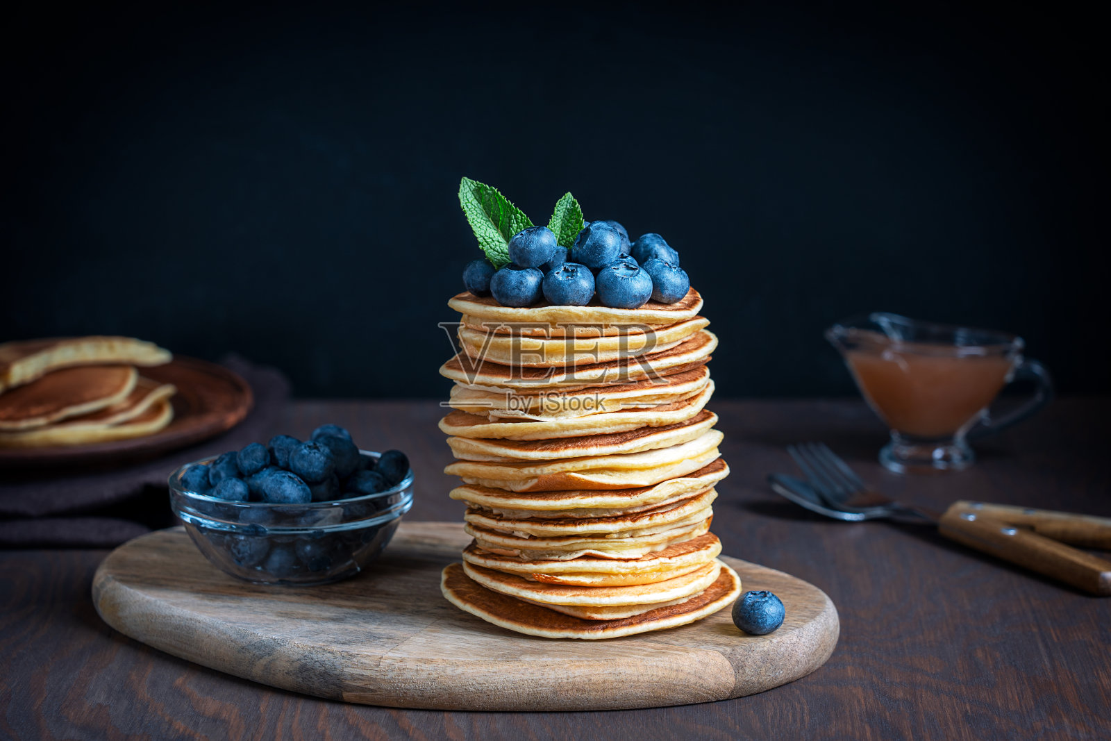 把自制的薄煎饼堆在新鲜的蓝莓和薄荷叶上,放在木砧板上,在深棕色的背景上放上成熟的浆果照片摄影图片