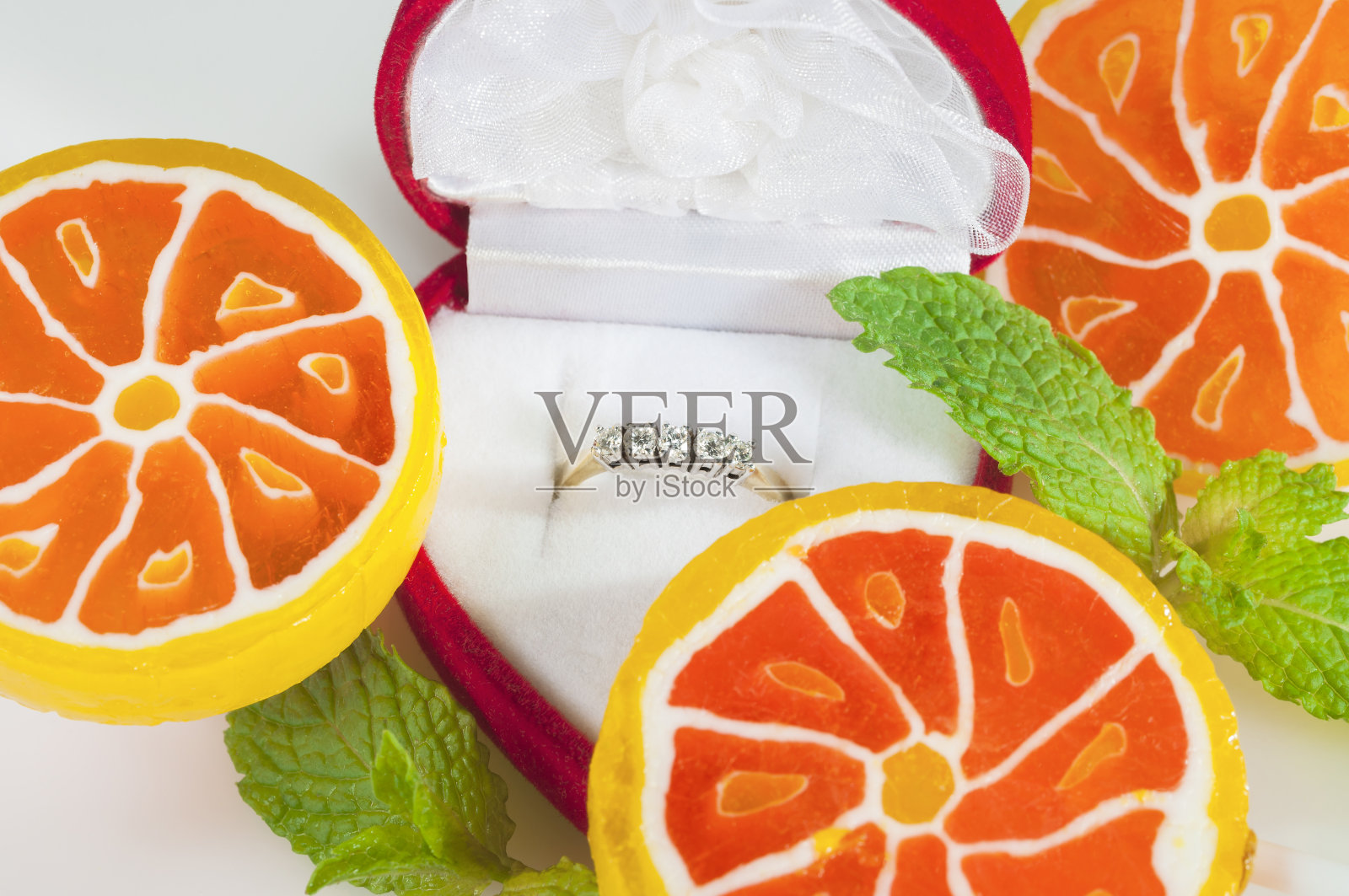 红色盒子里有钻石的戒指,周围是橙色糖果和新鲜薄荷叶。照片摄影图片