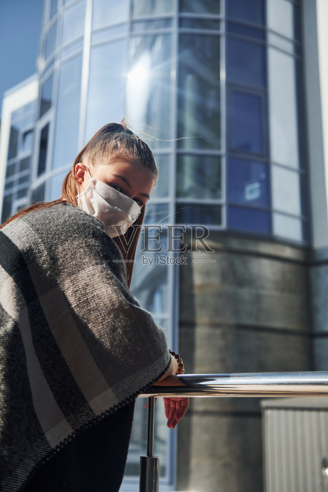 在阳光明媚的白天,年轻女子戴着防护面具站在商业大厦附近的户外。冠状病毒的概念照片摄影图片