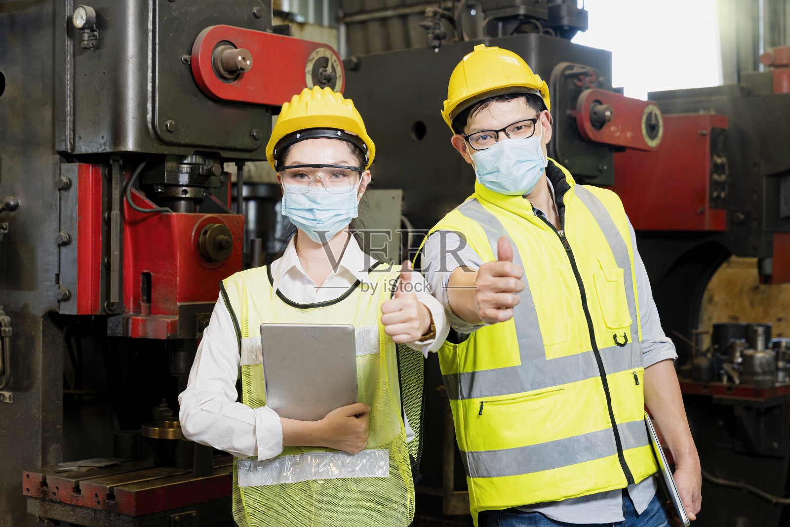 亚洲女人和男人戴着面具,拿着平板电脑,在一个工业工厂里竖起大拇指。在工厂里,专业技术人员正拿着平板电脑来控制工作。照片摄影图片