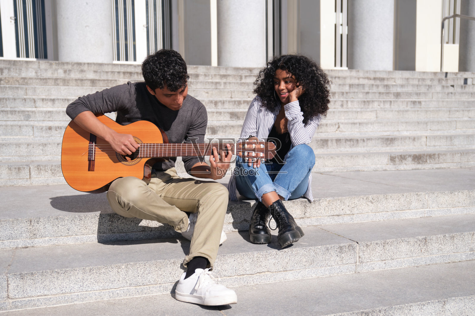 一对年轻的拉丁夫妇坐在楼梯上,弹着吉他,唱着歌。千禧世代。照片摄影图片