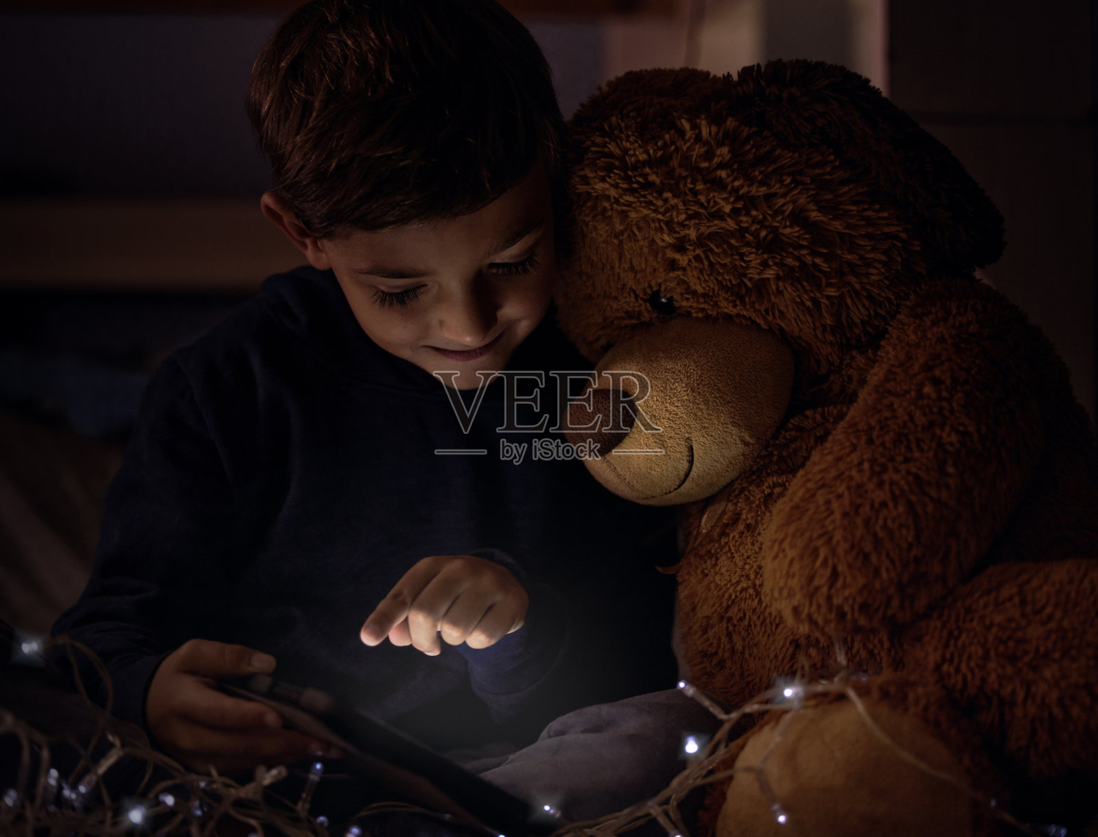 可爱的小男孩晚上在卧室里和泰迪熊一起使用平板电脑照片摄影图片