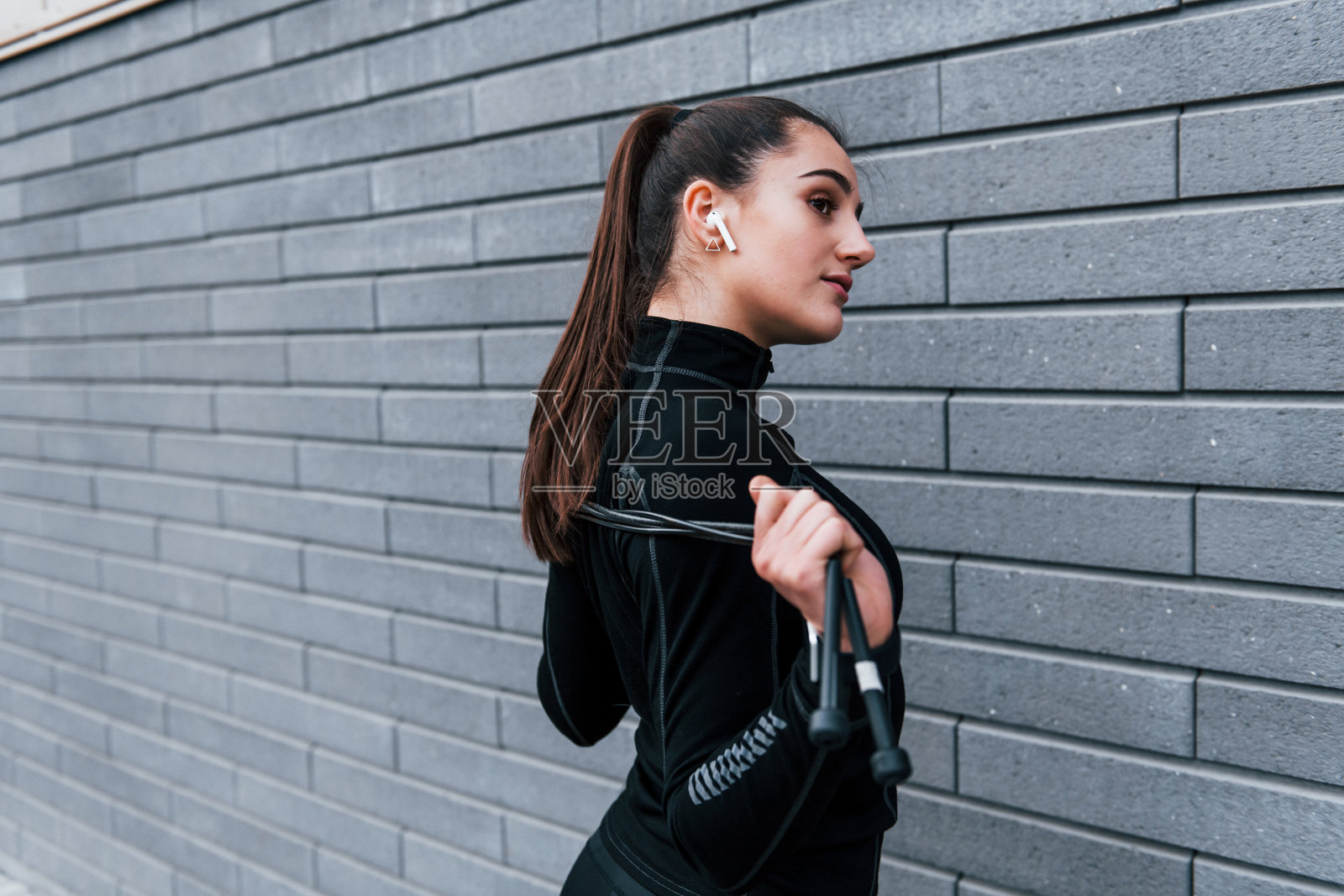 年轻的运动女孩站在黑色运动服与跳绳在户外附近的灰色墙壁照片摄影图片